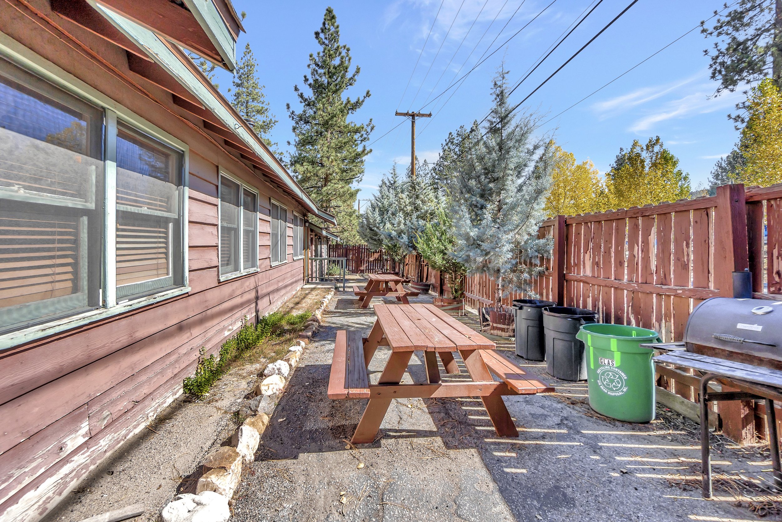 Backyard scene with a red wooden house, wooden picnic tables, green trash bins, a wooden fence, tall trees, power lines, and a clear blue sky.