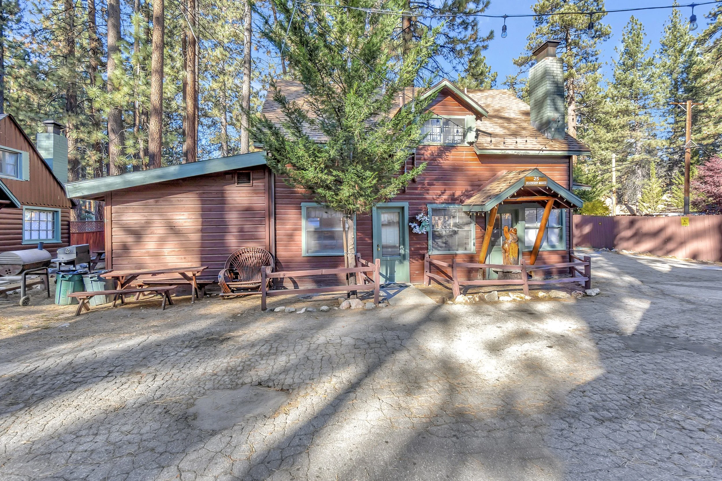 A rustic wooden house surrounded by tall pine trees, with a string of outdoor lights hanging overhead. In front of the house, there is outdoor seating including a bench and chairs, a small table, and some outdoor accessories, on a cracked concrete or