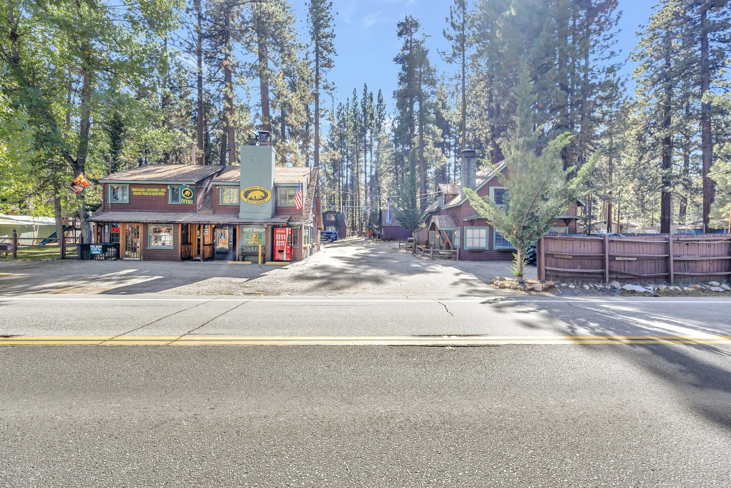 A small rustic shop with a red wooden exterior, signs, and vending machines, set among tall pine trees in a forested area, with a dirt path and a parking space in front.