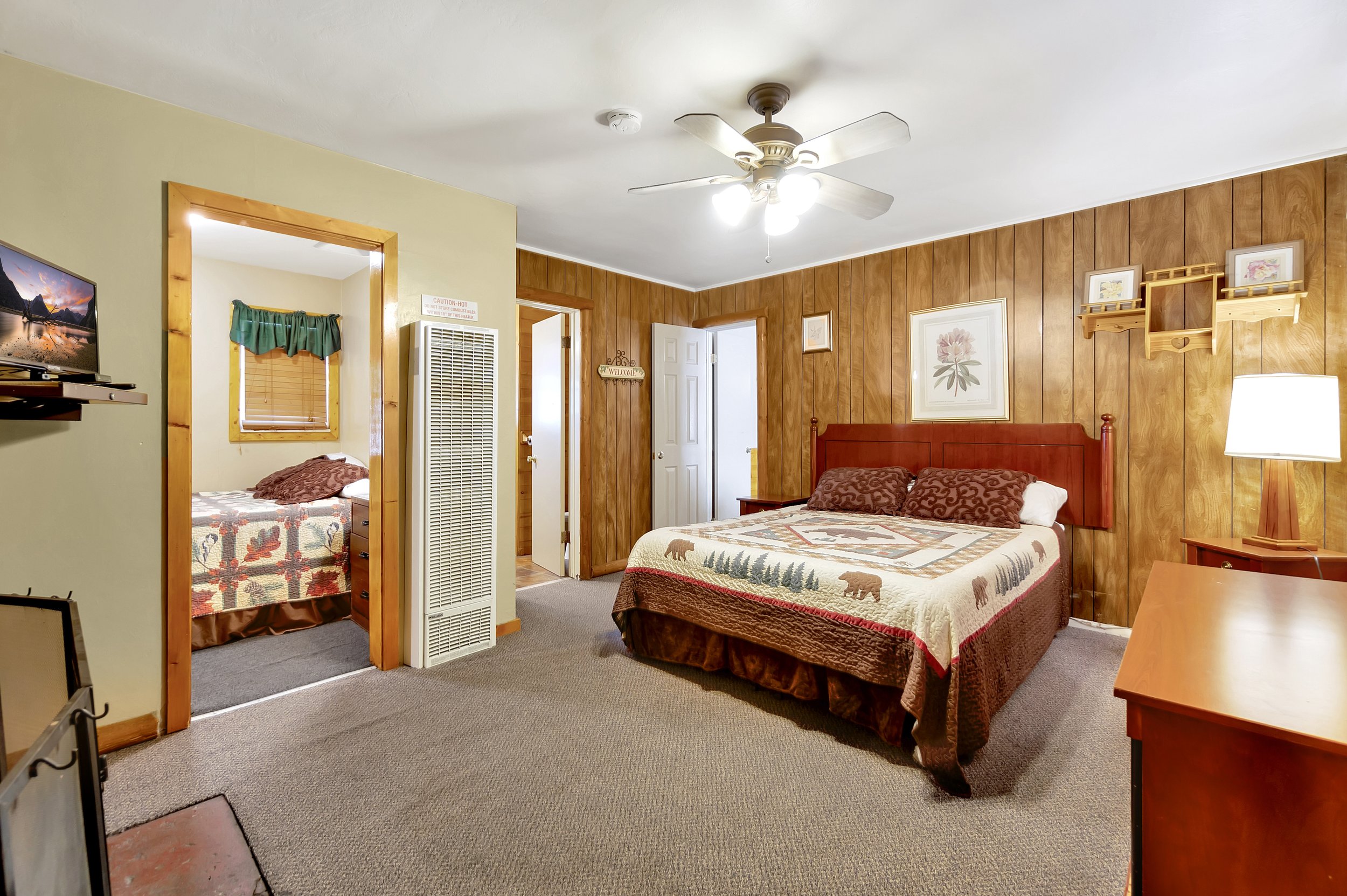 Interior view of a bedroom with wood-paneled walls, a ceiling fan with lights, a framed flower artwork, a wooden bed with a bear-themed quilt, a bedside lamp, and a small table. Partially visible are a small adjacent bedroom with a bed and a window w