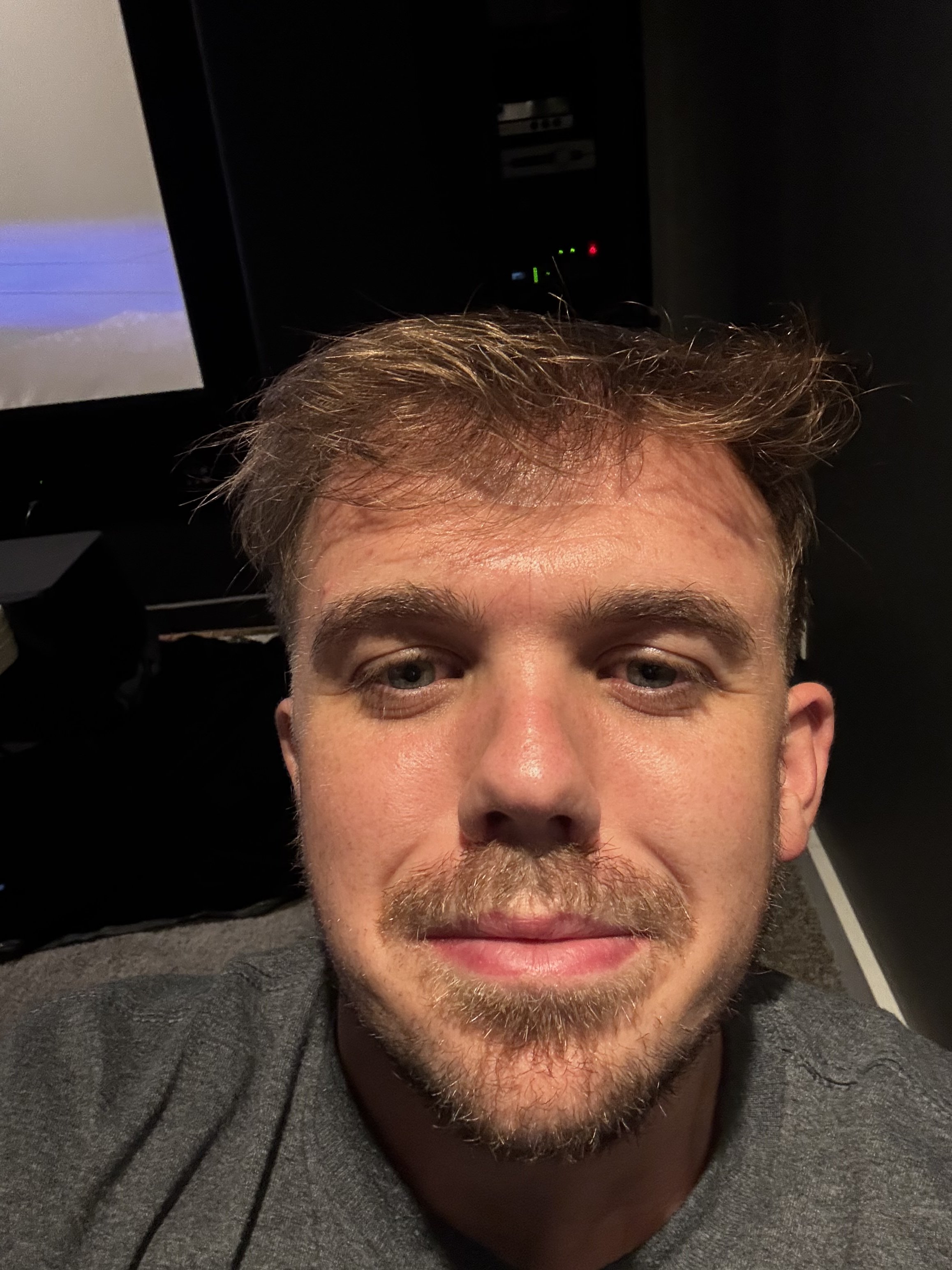 A close-up of a man's face, showing his eyes, nose, and mouth, with short light brown hair, some facial hair, and wearing a gray shirt, taken at a slight angle from above.