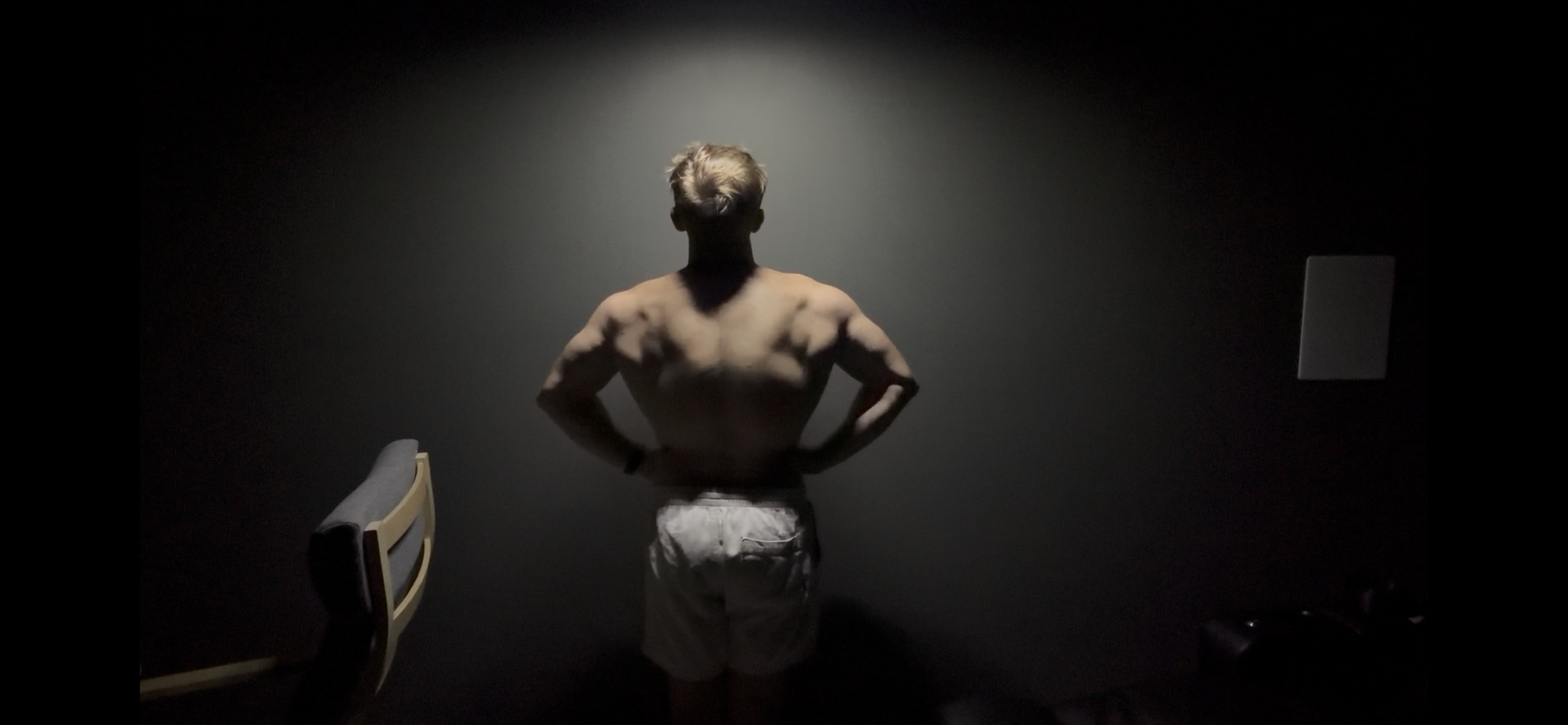 A shirtless man with light hair viewed from behind in a dimly lit room, standing with hands on hips, facing a dark wall.