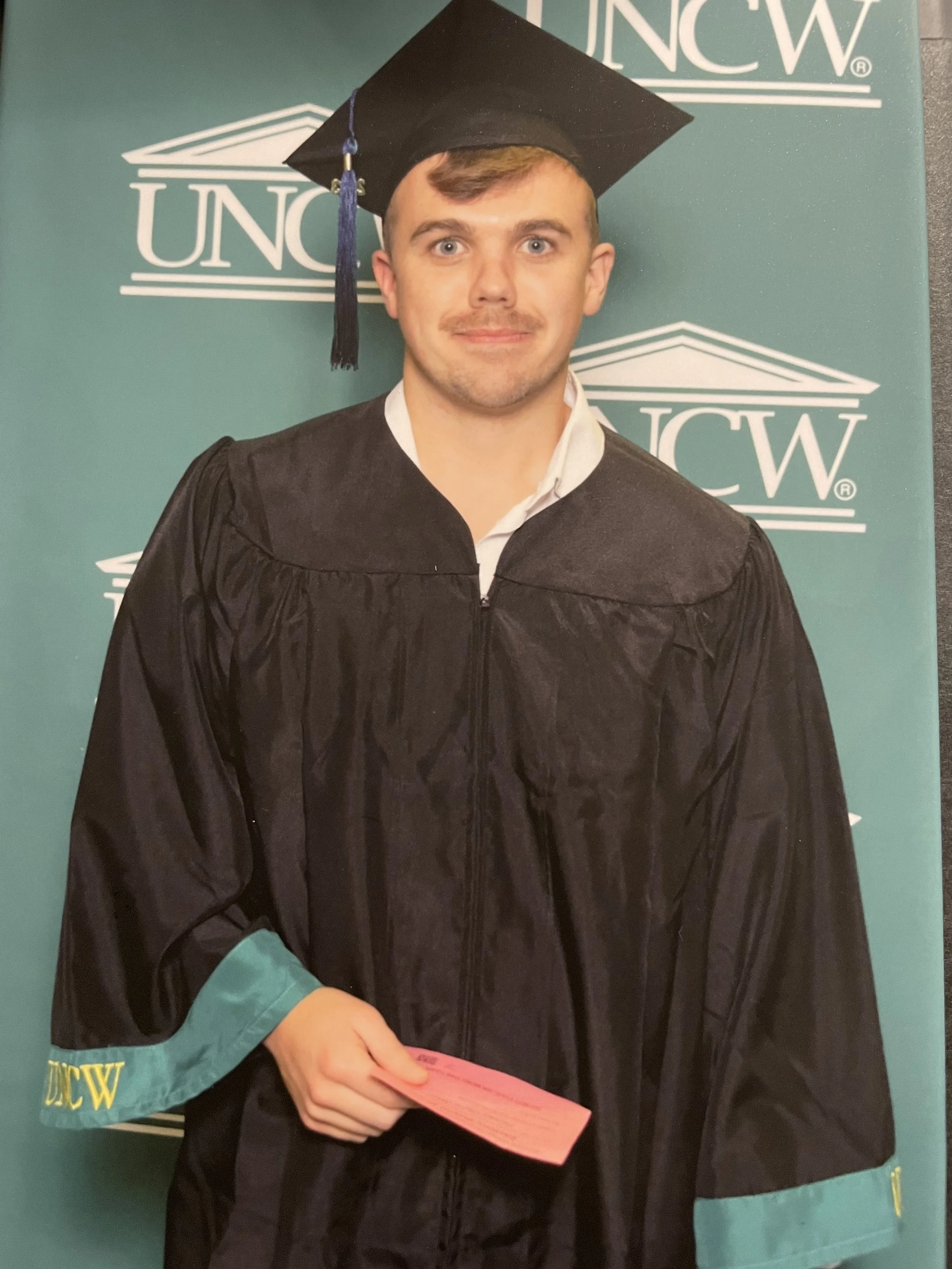Young man in graduation cap and gown holding a pink diploma, standing in front of a green background with 'UNCW' logo.