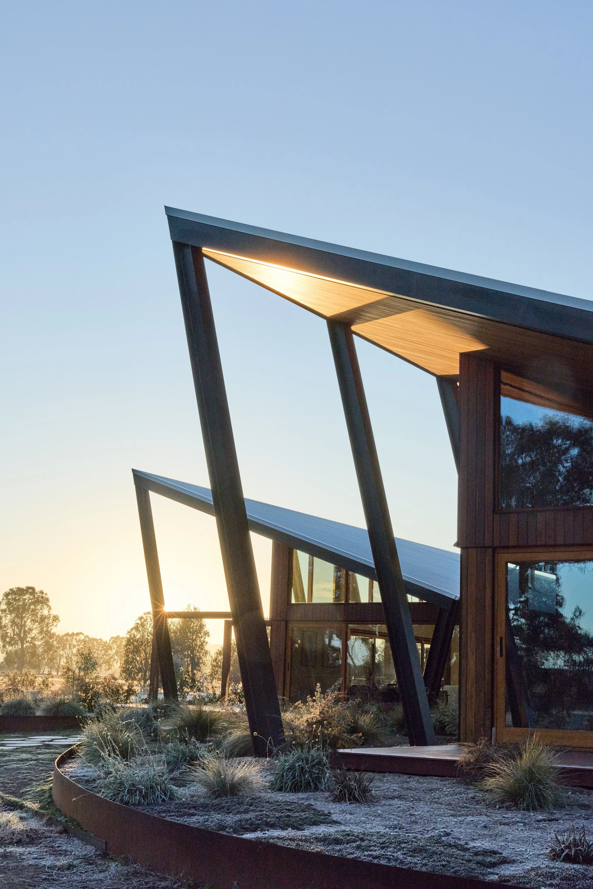 Modern house with large overhanging roof supported by angled columns, large windows, and built with wood and metal materials. The house is set against a sunset sky, with trees in the background and a garden with drought-tolerant plants in the foreground.