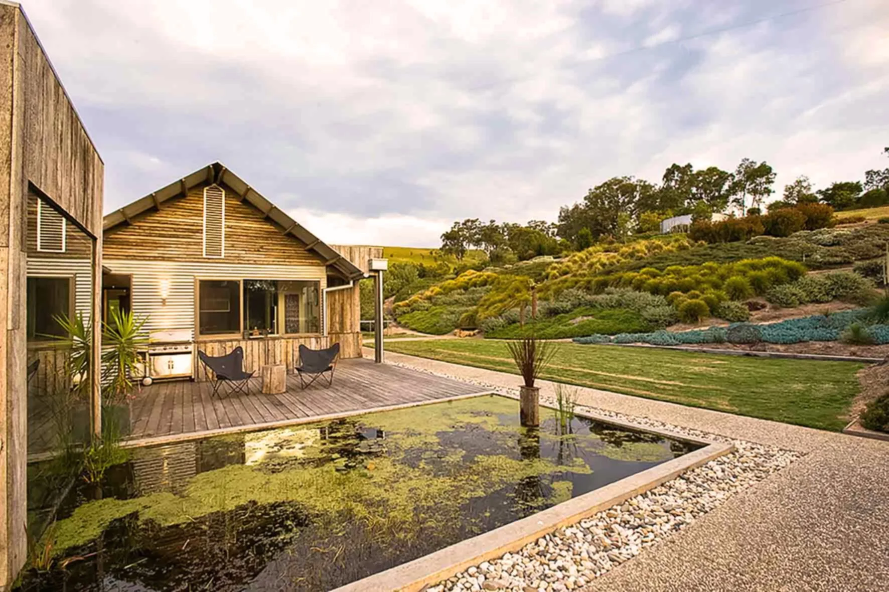 A modern house with wooden exterior, wooden deck with two chairs, a pond with green algae, and a landscaped garden with hills and trees in the background under a cloudy sky.