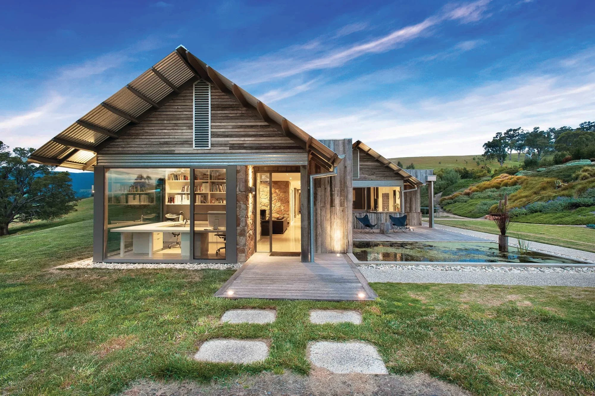 A modern house with large glass windows, a sloped roof, and outdoor seating, set in a green landscape with trees and hills in the background during evening or dusk.