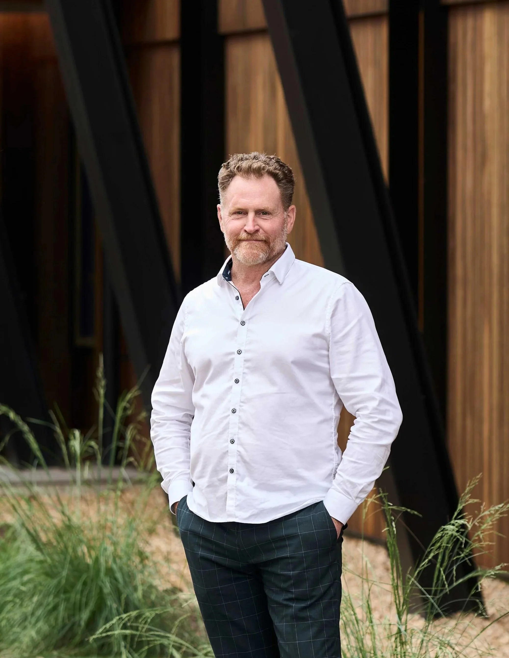 A man with light skin, a beard, and short curly hair, wearing a white button-up shirt and dark plaid pants, standing outdoors in front of a building with wooden panels and black structural elements, with plants in the foreground.
