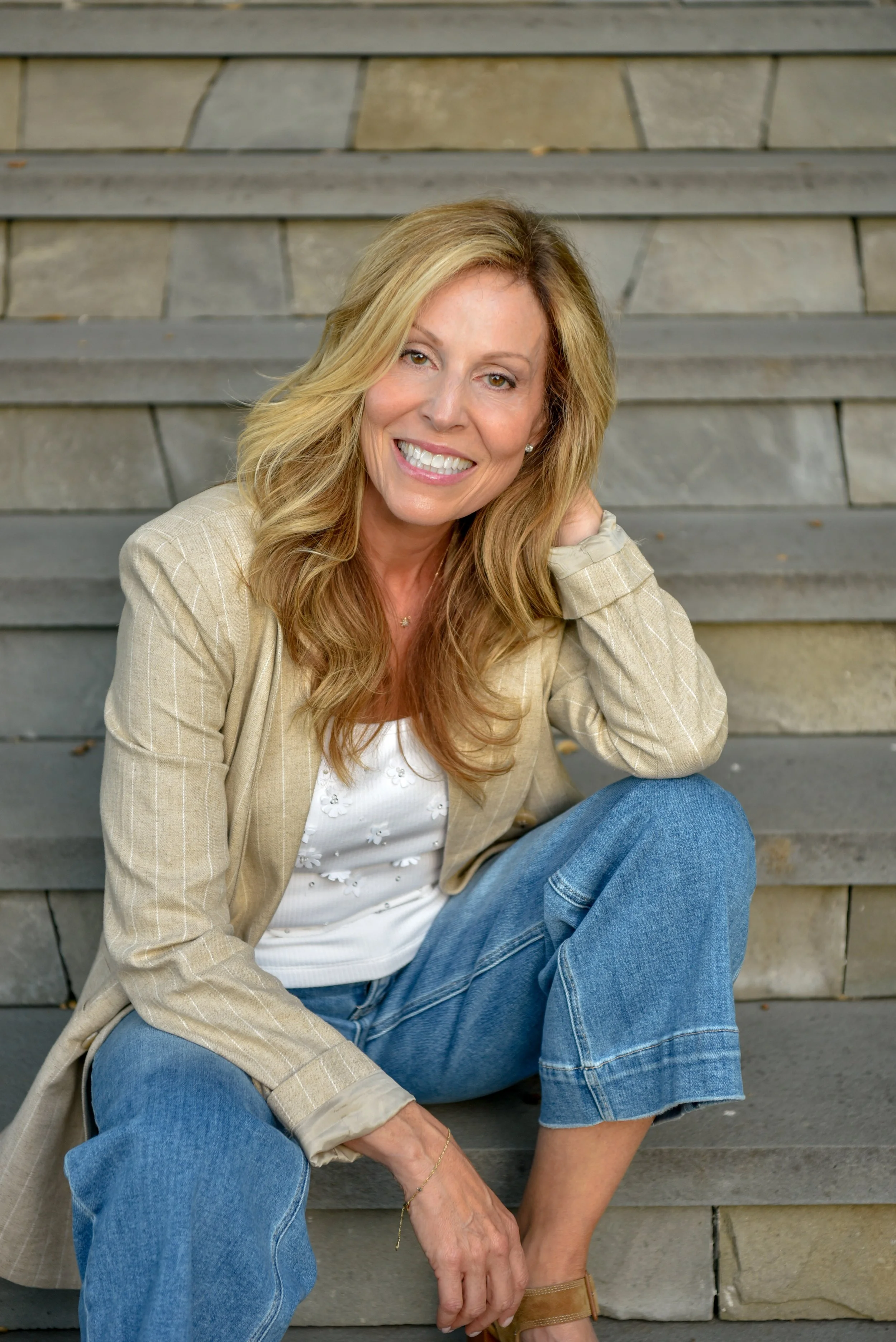 A woman with long blonde hair smiling, sitting on wooden stairs, wearing a beige blazer, white blouse, blue jeans, and tan shoes.