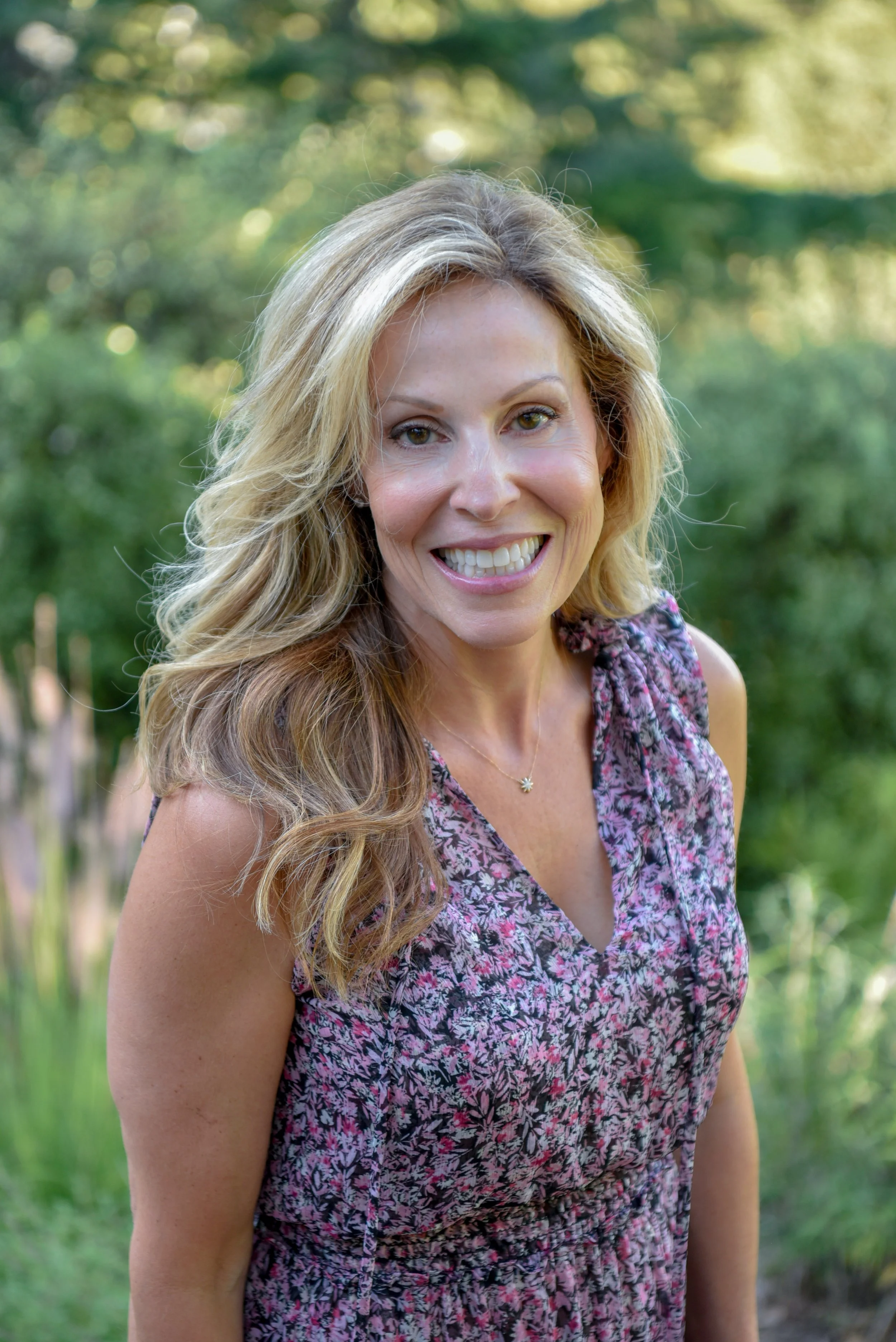 A woman with long wavy blonde hair smiling outdoors with a blurred green foliage background.