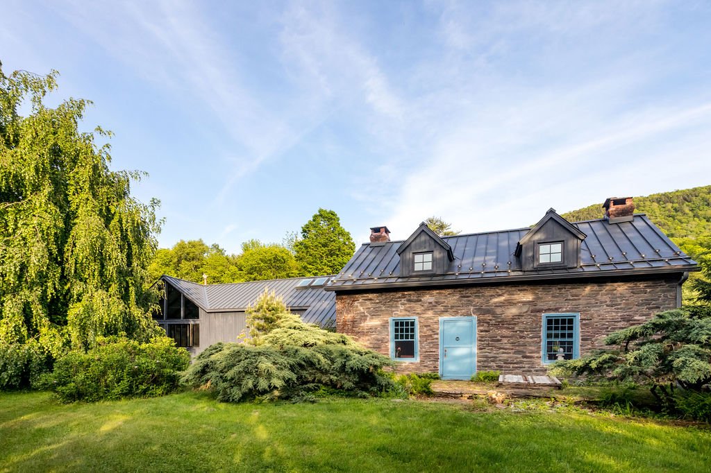 A stone house with a blue window and door, surrounded by lush greenery and trees under a partly cloudy sky.