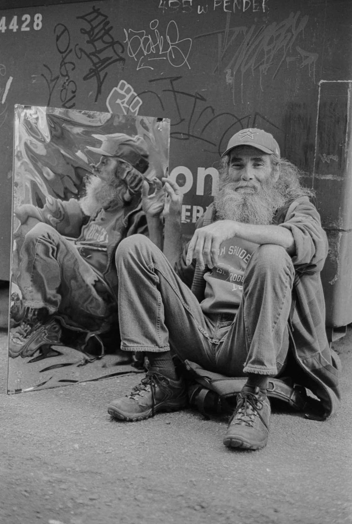 A homeless man with a long beard, wearing a cap and casual clothing, sitting on the ground against a graffitied wall, next to a portrait of an older man with a beard, holding a pipe and wearing a cap.