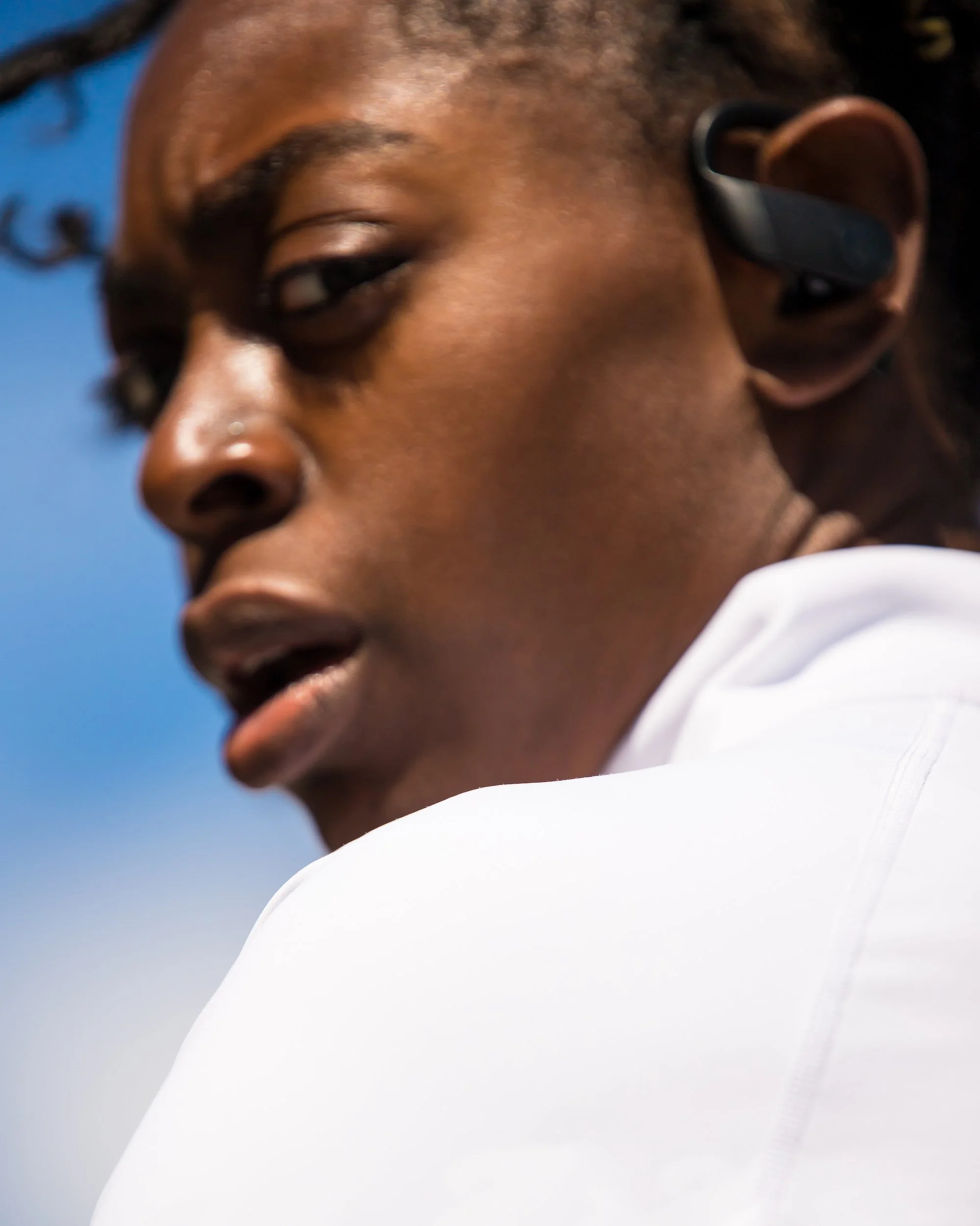 Close-up of a person's face, showing the right eye, nose, and cheek, with an earbud in the right ear, against a blue sky background.