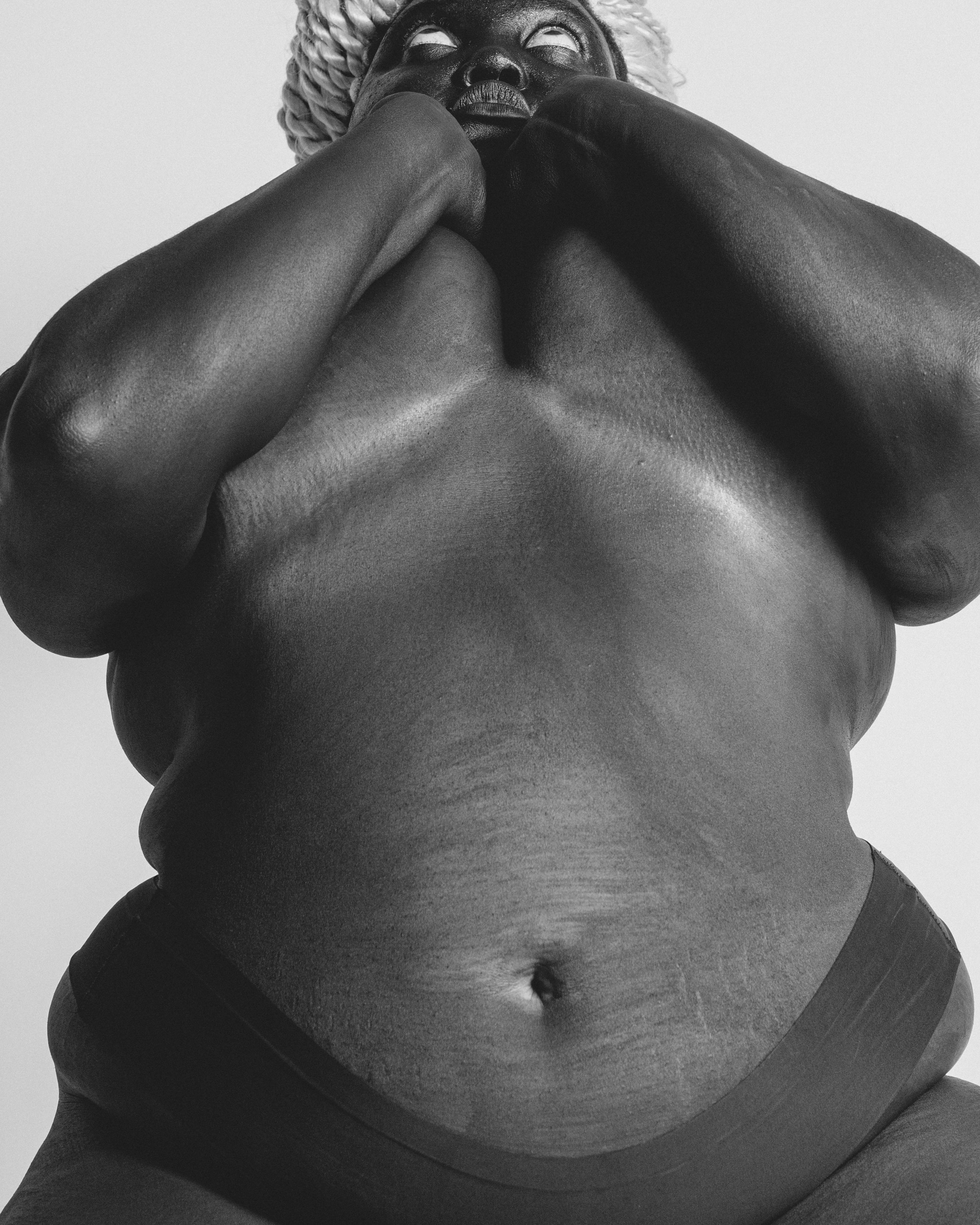 Black and white close-up photograph of a person's chest, neck, and chin with the person's face partially visible. The person is looking upwards, with their hands resting on their face, creating a contemplative pose. The image captures the skin textur