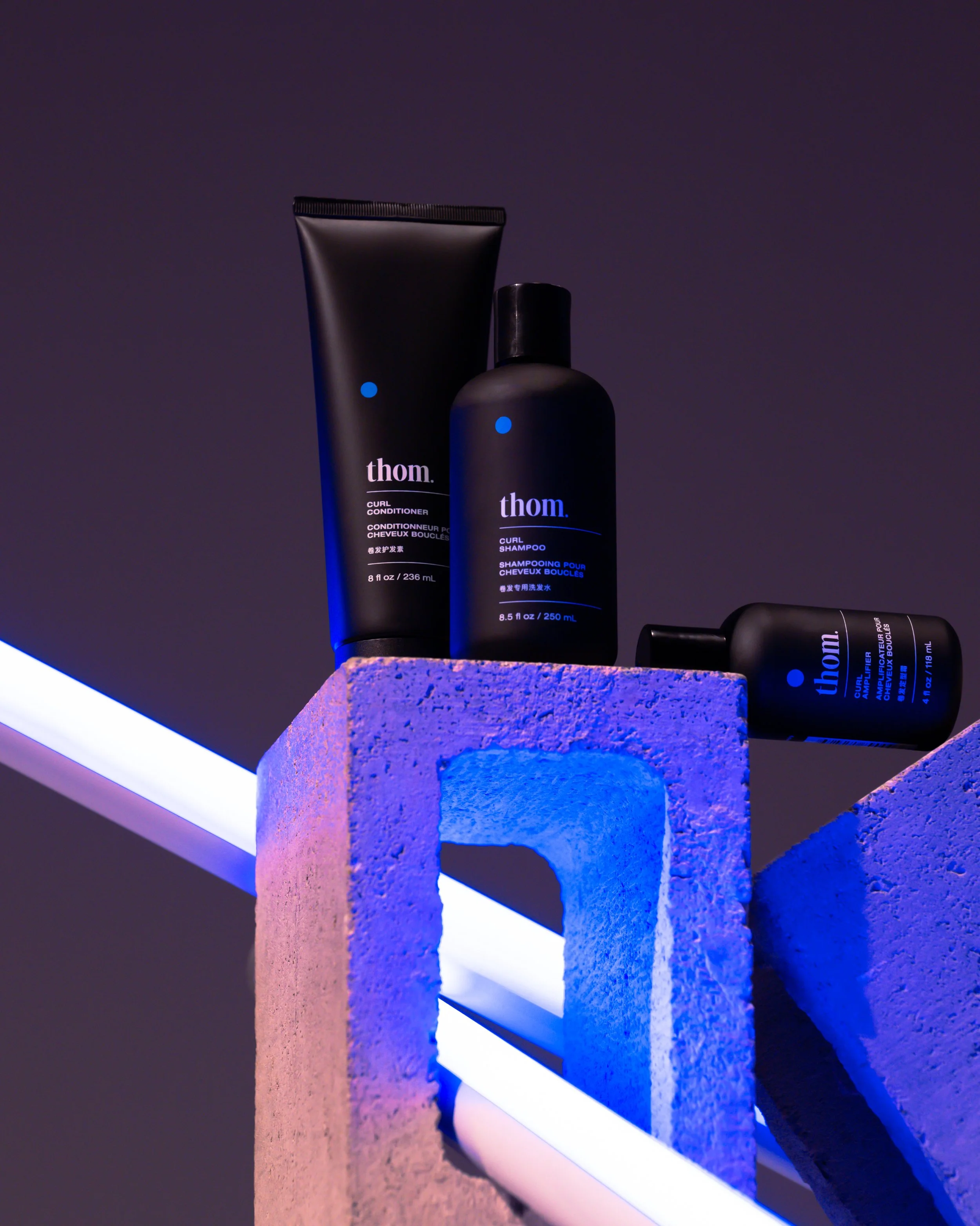 Two black bottles of Thomm hair care products on a textured blue and pink pedestal against a dark background with colored lighting, one labeled curl conditioner and the other curl shampoo.