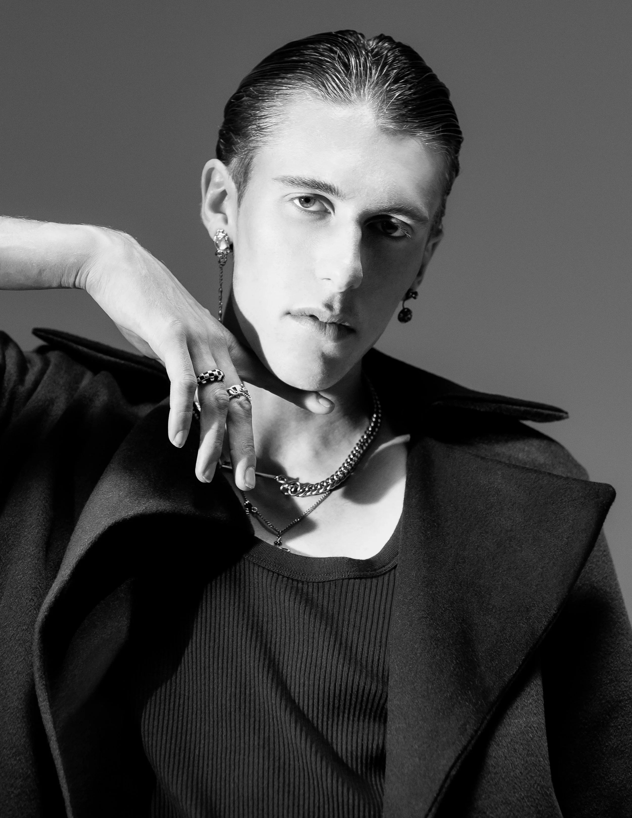 A black and white portrait of a young woman with slicked-back hair, wearing earrings, layered necklaces, and rings, with one hand raised near the face, dressed in a zip-up jacket over a ribbed top.