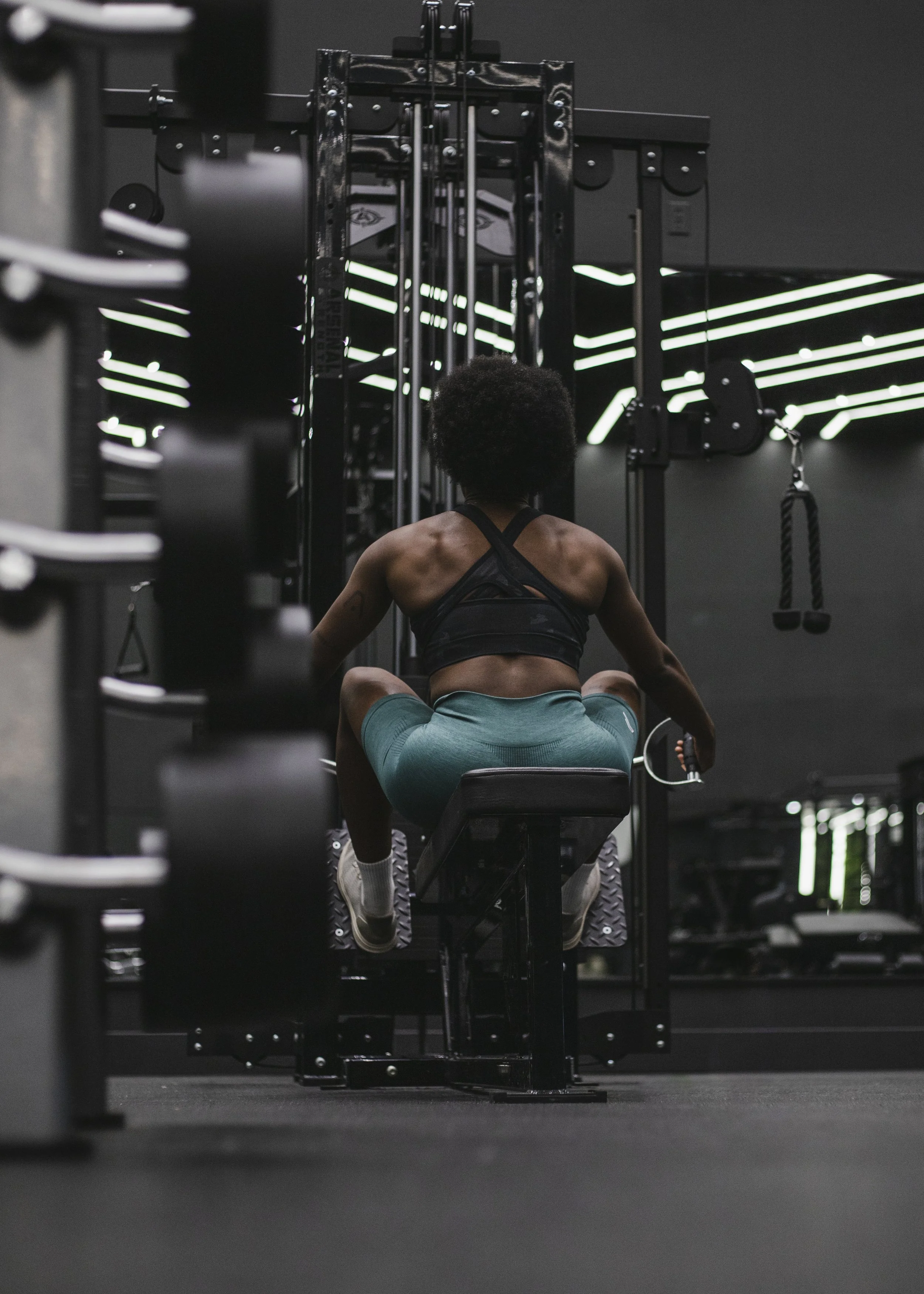 A person with curly hair wearing a black sports bra and teal shorts, sitting on a workout bench in a gym, facing a piece of fitness equipment.