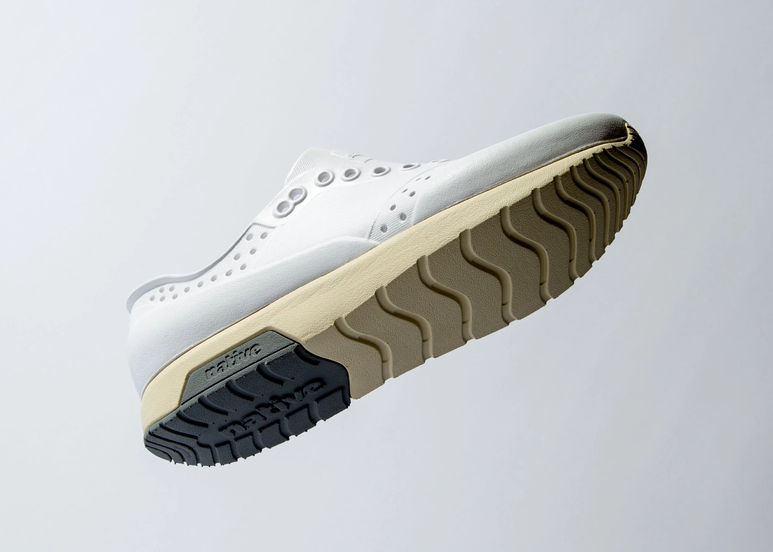 Close-up of a white sneaker with a beige midsole and black outsole, showing the tread pattern on the bottom.