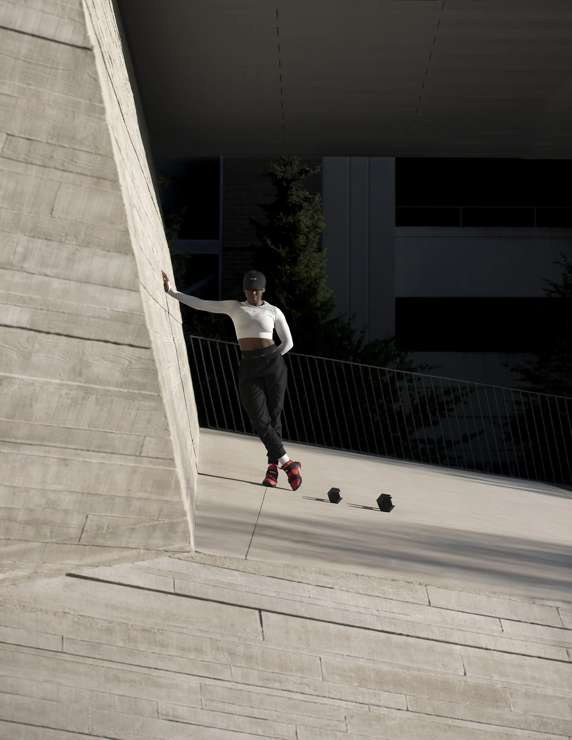 A person in athletic clothing and a baseball cap leaning against a low wall in an outdoor urban setting, with workout weights and a metal railing nearby.
