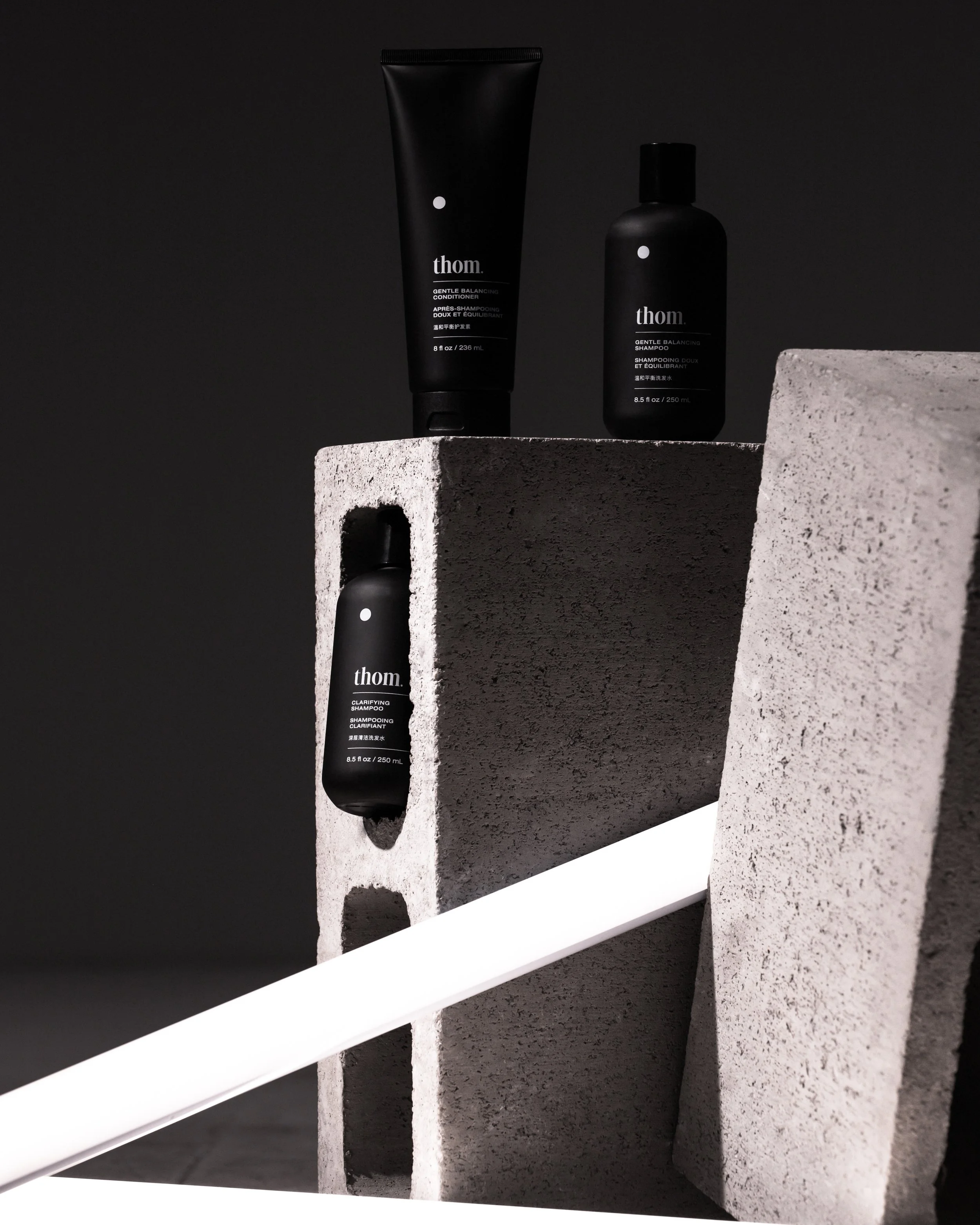 Black bottles of Thom hair care products, a gentle balancing conditioner and shampoo, displayed on concrete blocks against a black background.