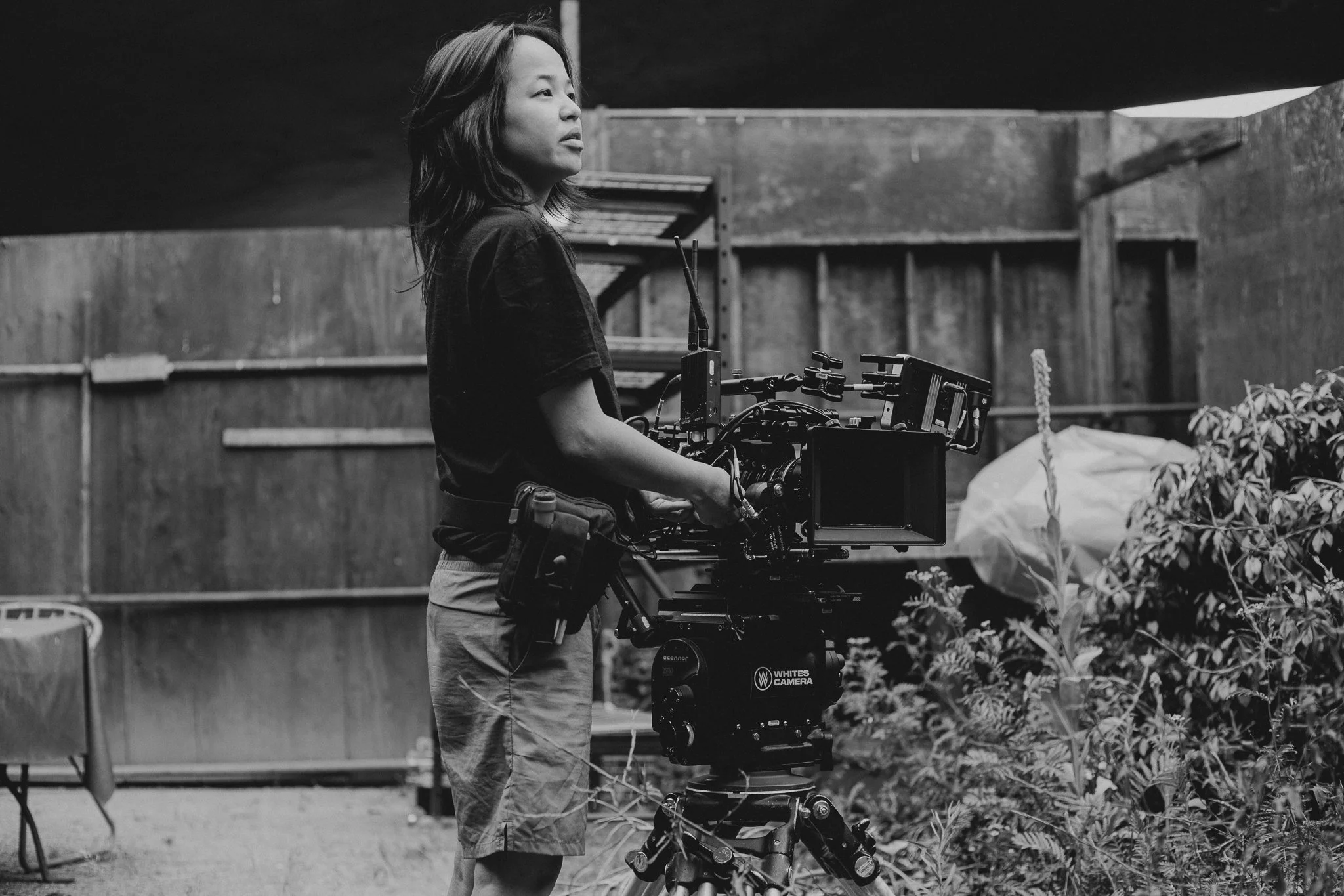 A woman operating a professional video camera on a tripod in an outdoor garden setting, with plants and wooden fencing around.