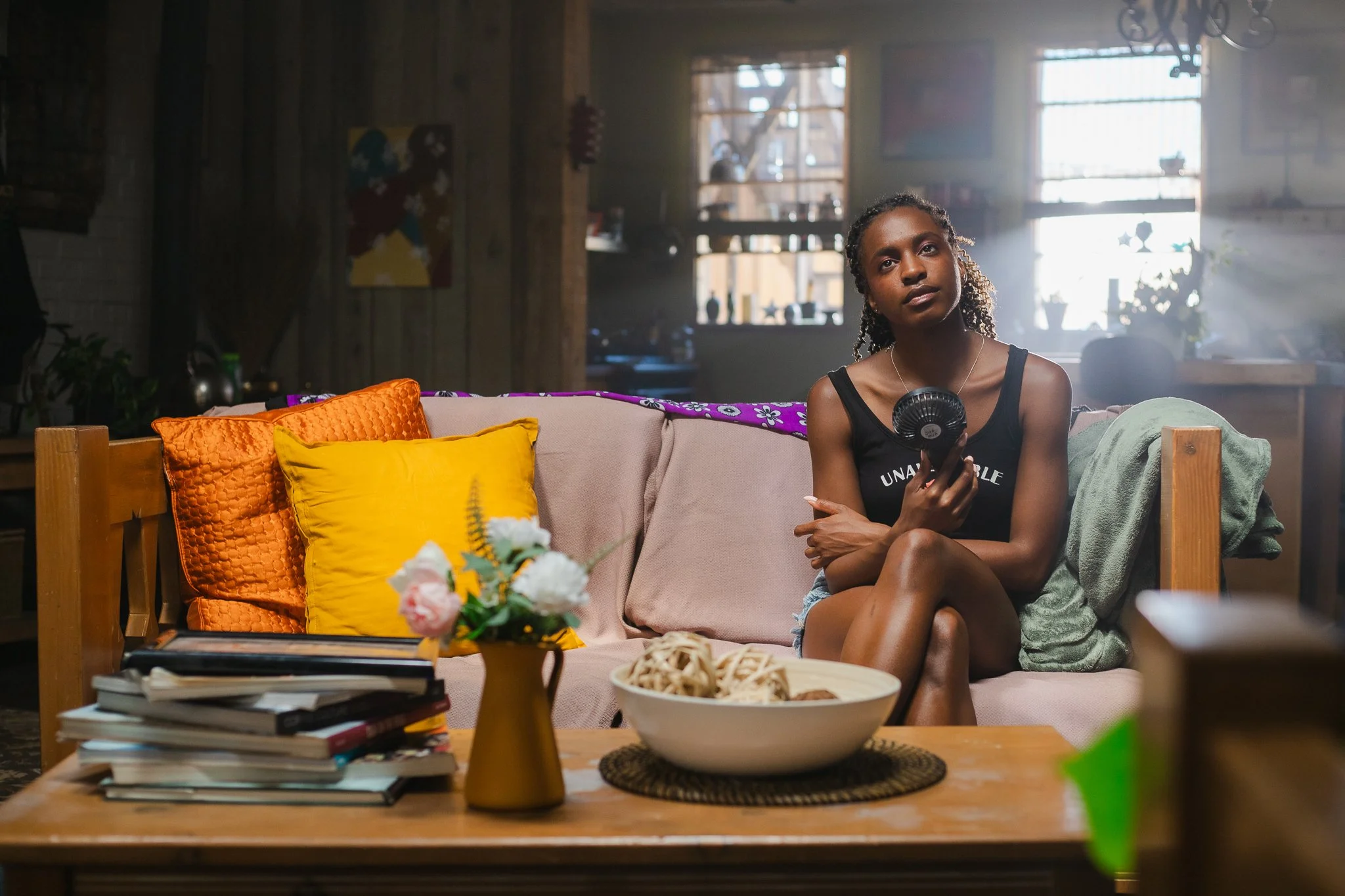 A young woman with curly hair in a ponytail sitting on a beige couch in a rustic living room, holding a small portable fan, with colorful pillows and a wooden table with magazines and a bowl of snacks in front of her. Sunlight filters through the win