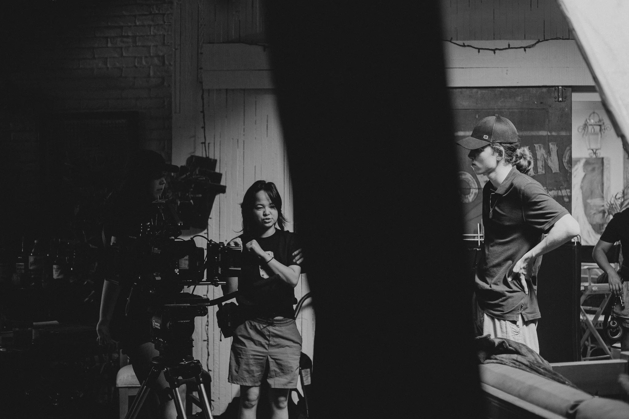 Black and white photograph of a film production set with a woman operating a camera, a young girl standing nearby, and a man with long hair and a cap standing with hands on hips. The background features wooden walls and hanging lights.