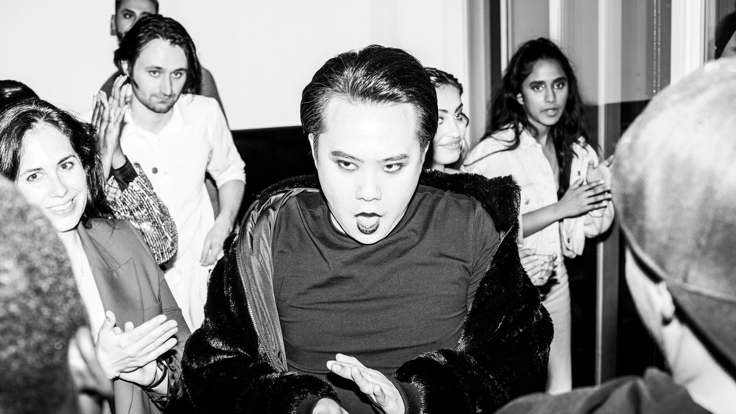 A black and white photo of a woman with short hair and dark lipstick, wearing a furry jacket, leaning forward with her mouth slightly open and hands together. Behind her, several people with various expressions are lined up, looking in her direction,