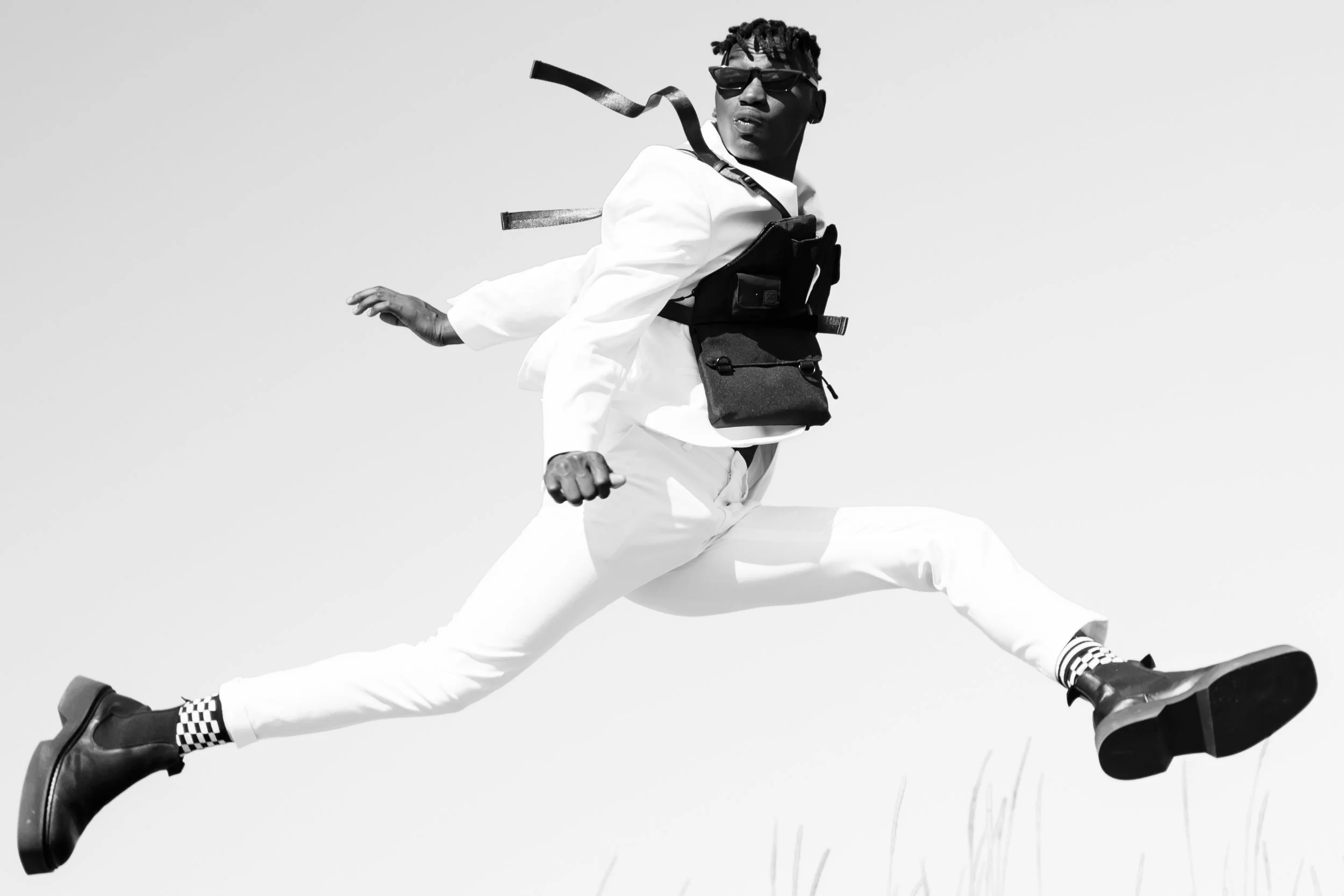 A person wearing sunglasses, a white outfit, and checkered socks, captured mid-jump against a plain background.