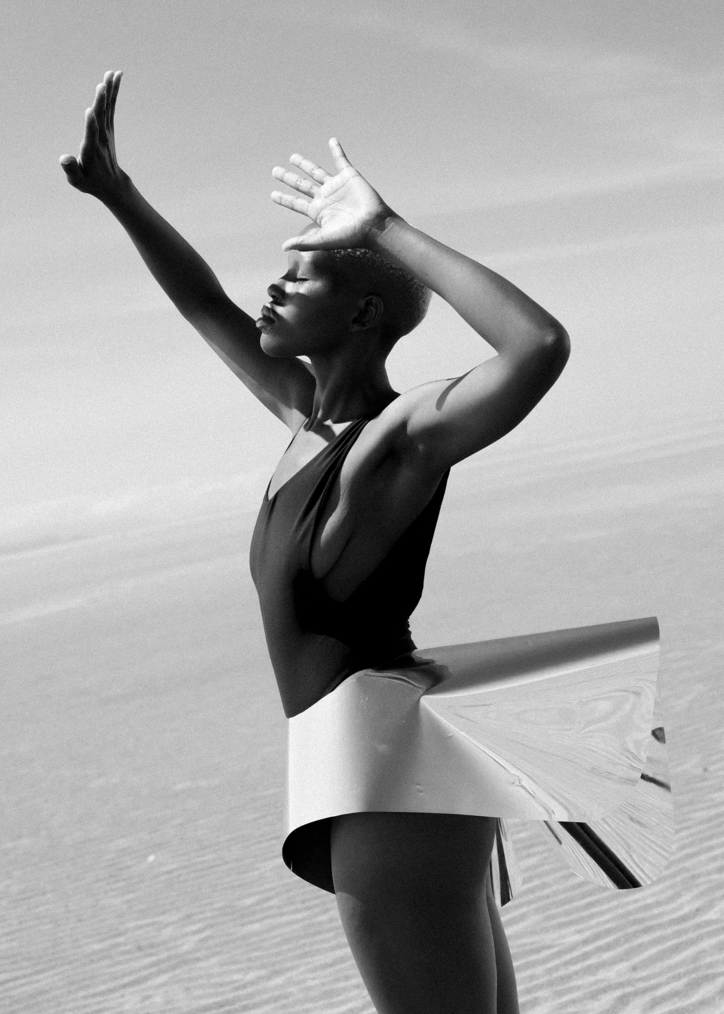 A woman standing on a beach with arms raised and eyes closed, wearing a swimsuit, in black and white
