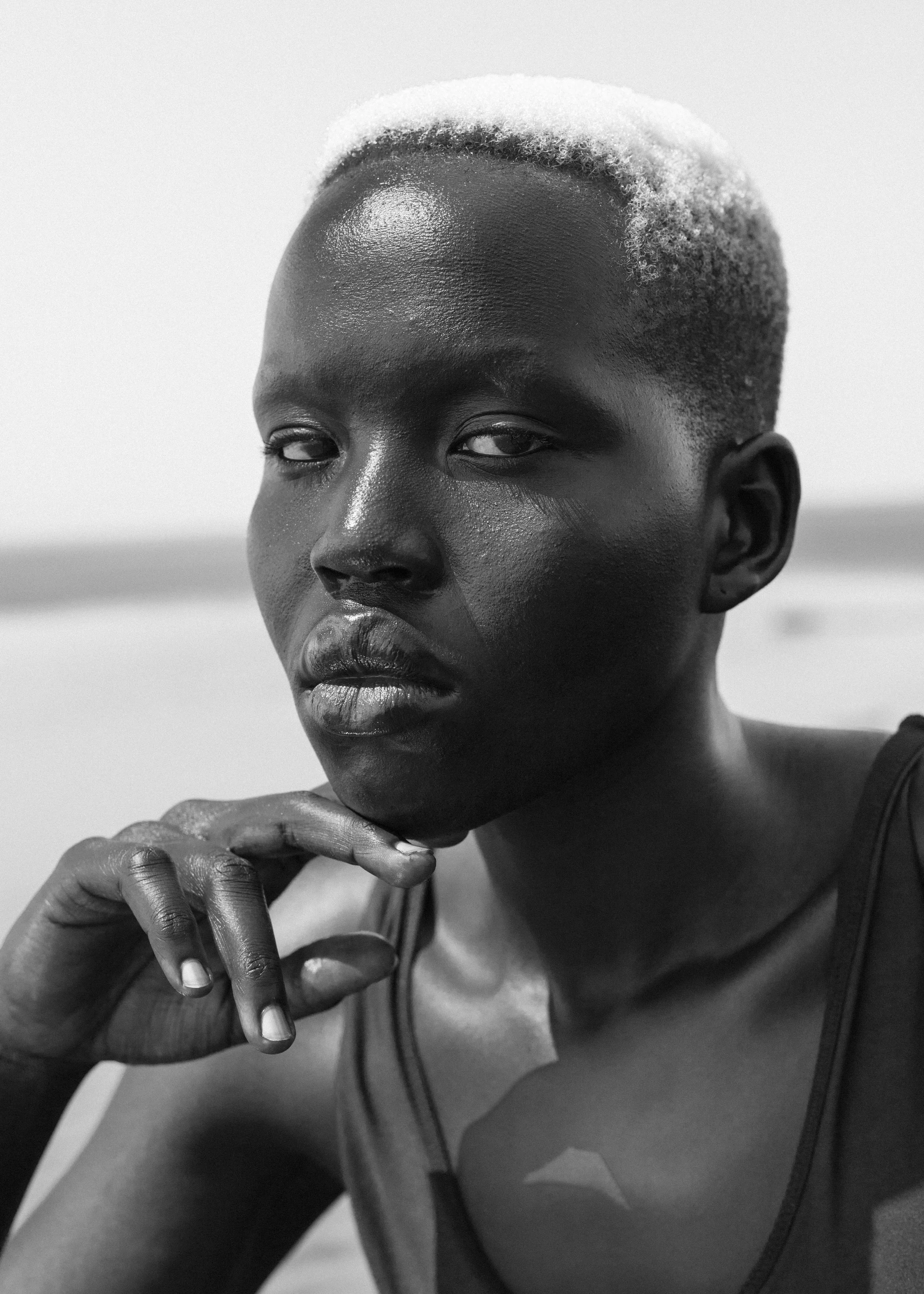 A close-up black and white photo of a person with short, light-colored hair, looking towards the camera with a serious expression. Their hand is resting near their chin, and they are wearing a sleeveless top.