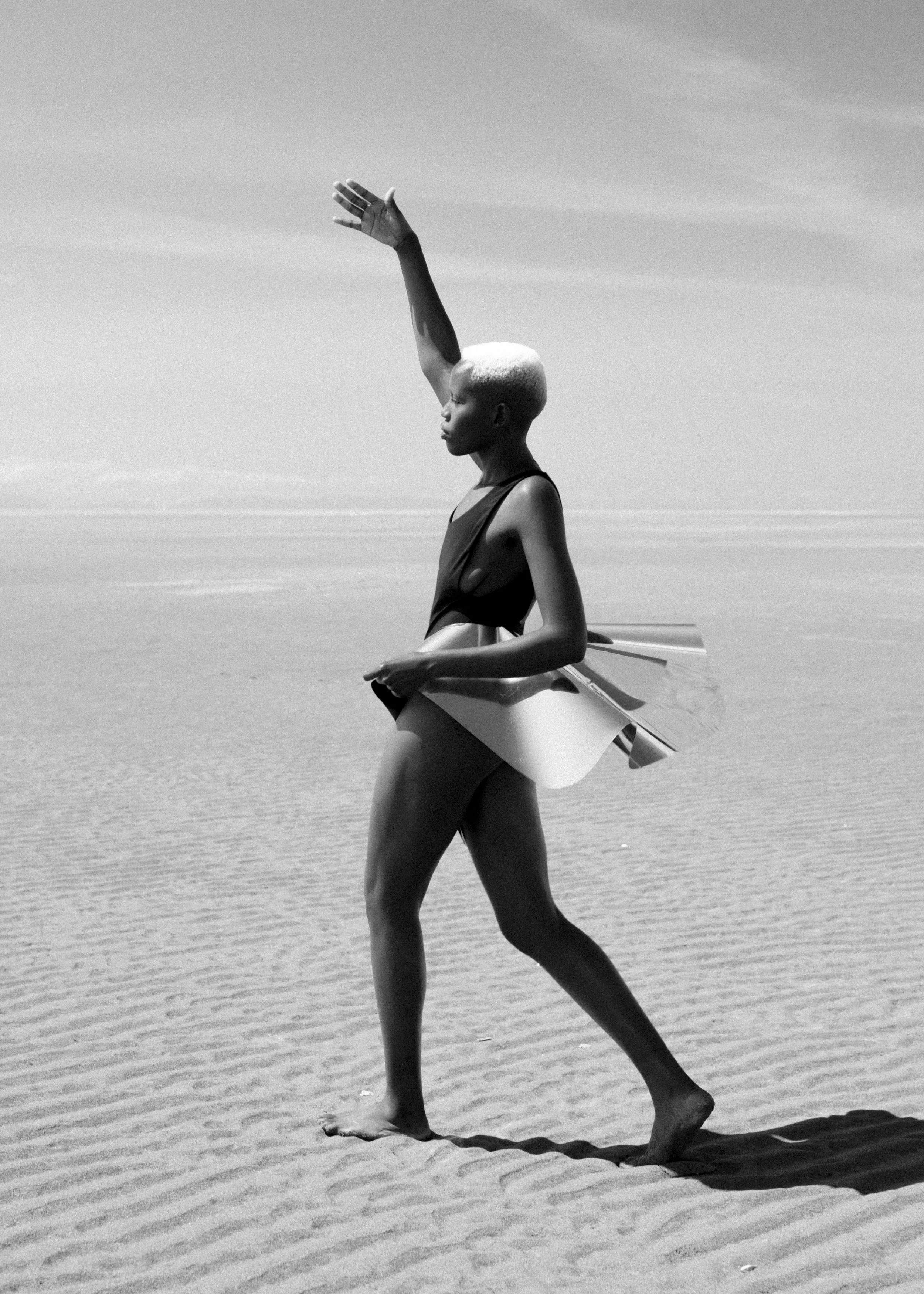 A woman with short light-colored hair walking on a sandy beach with a surfboard under her arm.