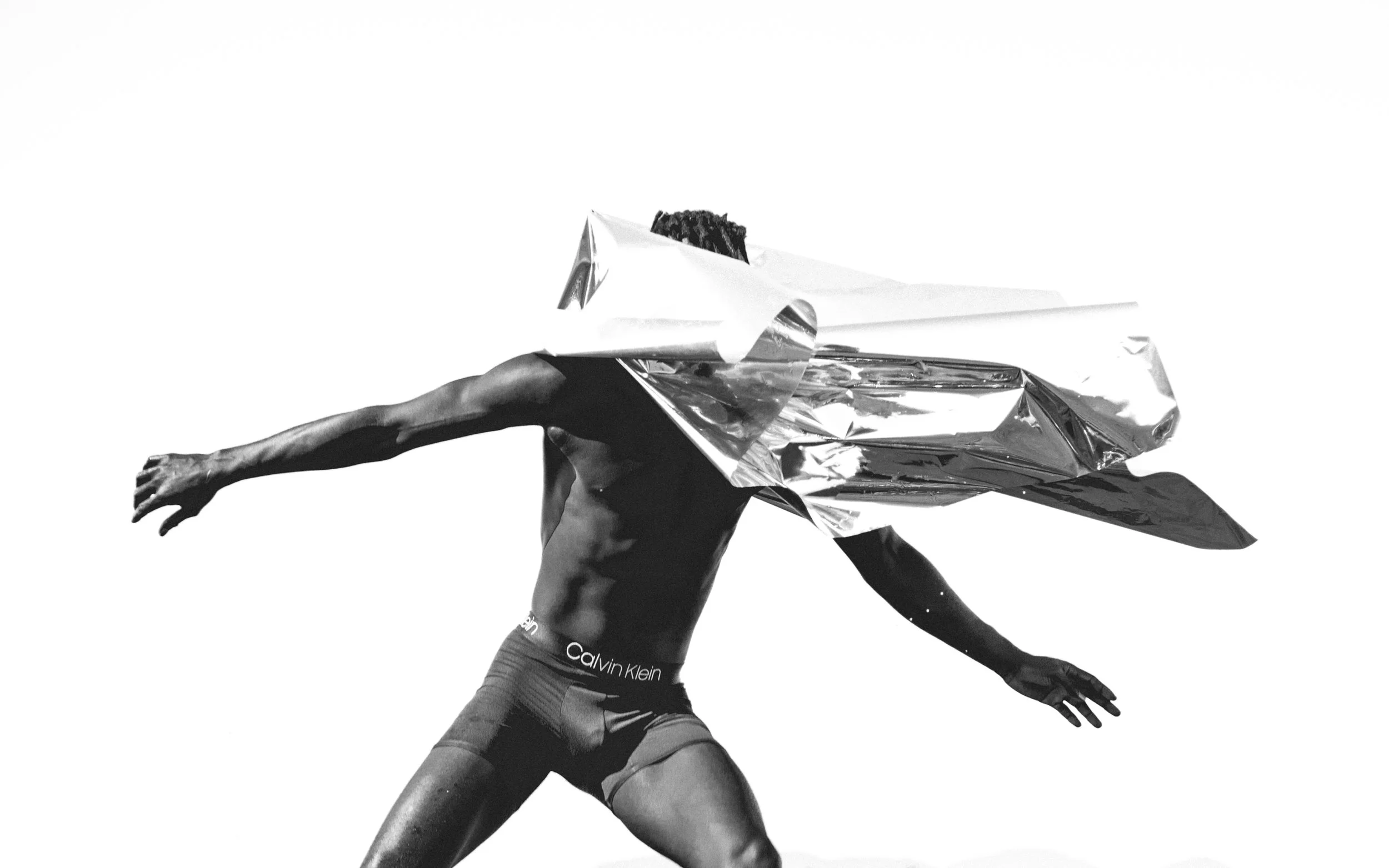 Black and white photo of a person in athletic shorts, with arms extended and wearing a metallic, reflective, geometrically folded shirt over their head.