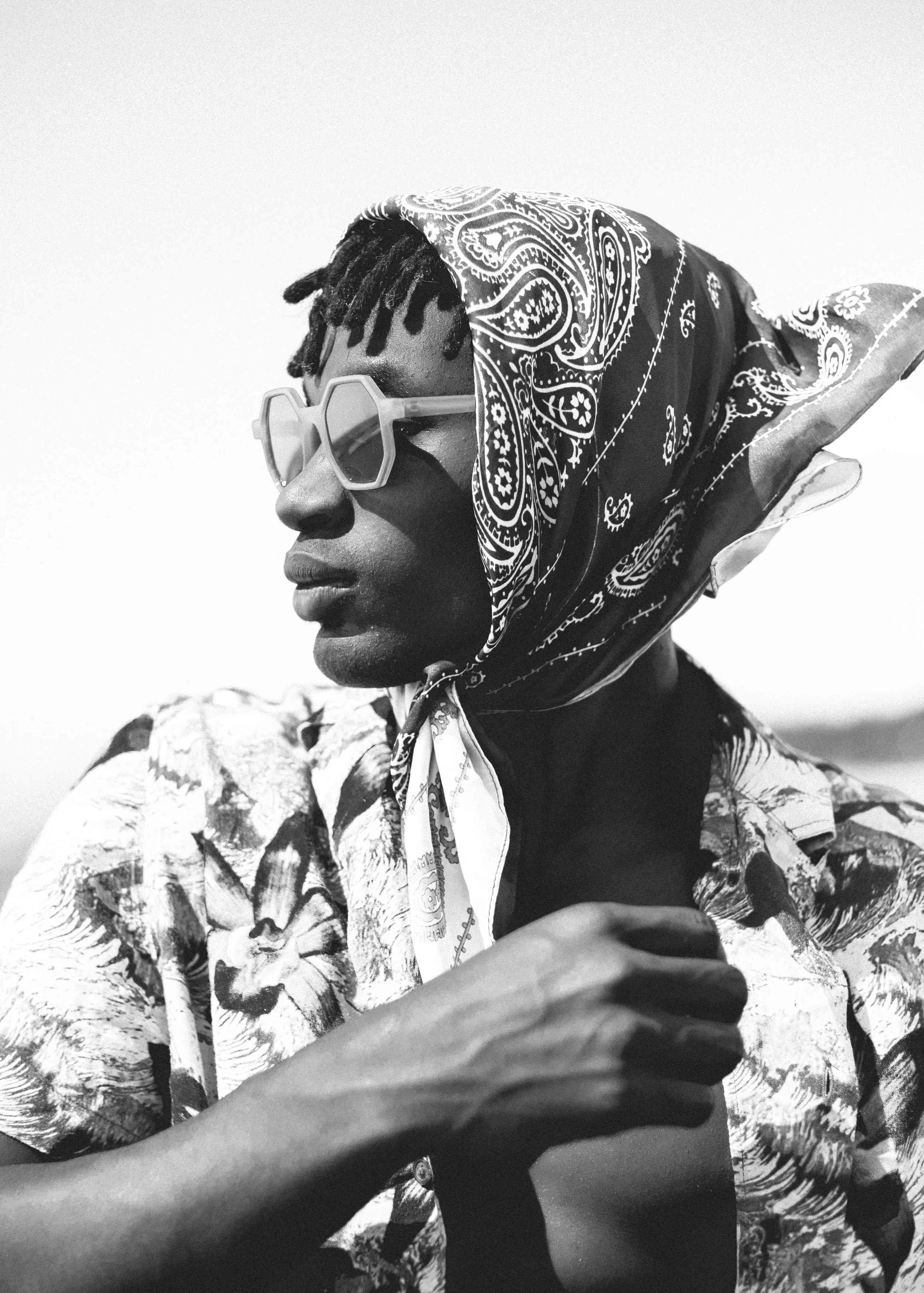 A black and white photo of a person, viewed in profile, wearing sunglasses, a patterned headscarf, and a floral shirt, with their arm crossed.