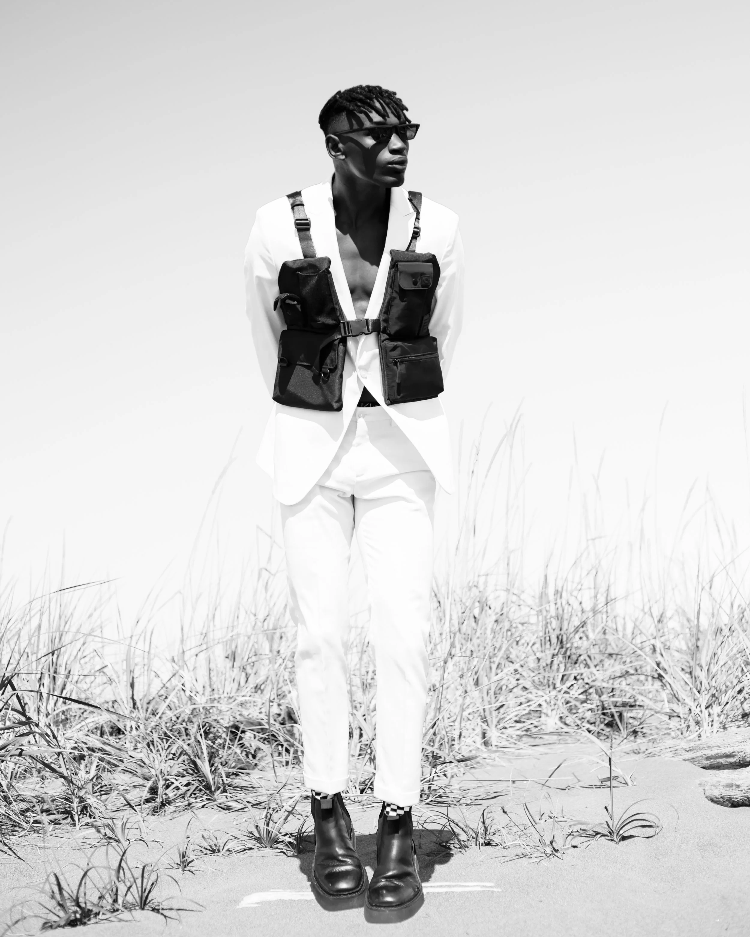 A person standing outdoors in a sunny, open area with tall grass, wearing sunglasses, a white shirt, a tactical vest, white pants, checkered socks, and black boots.