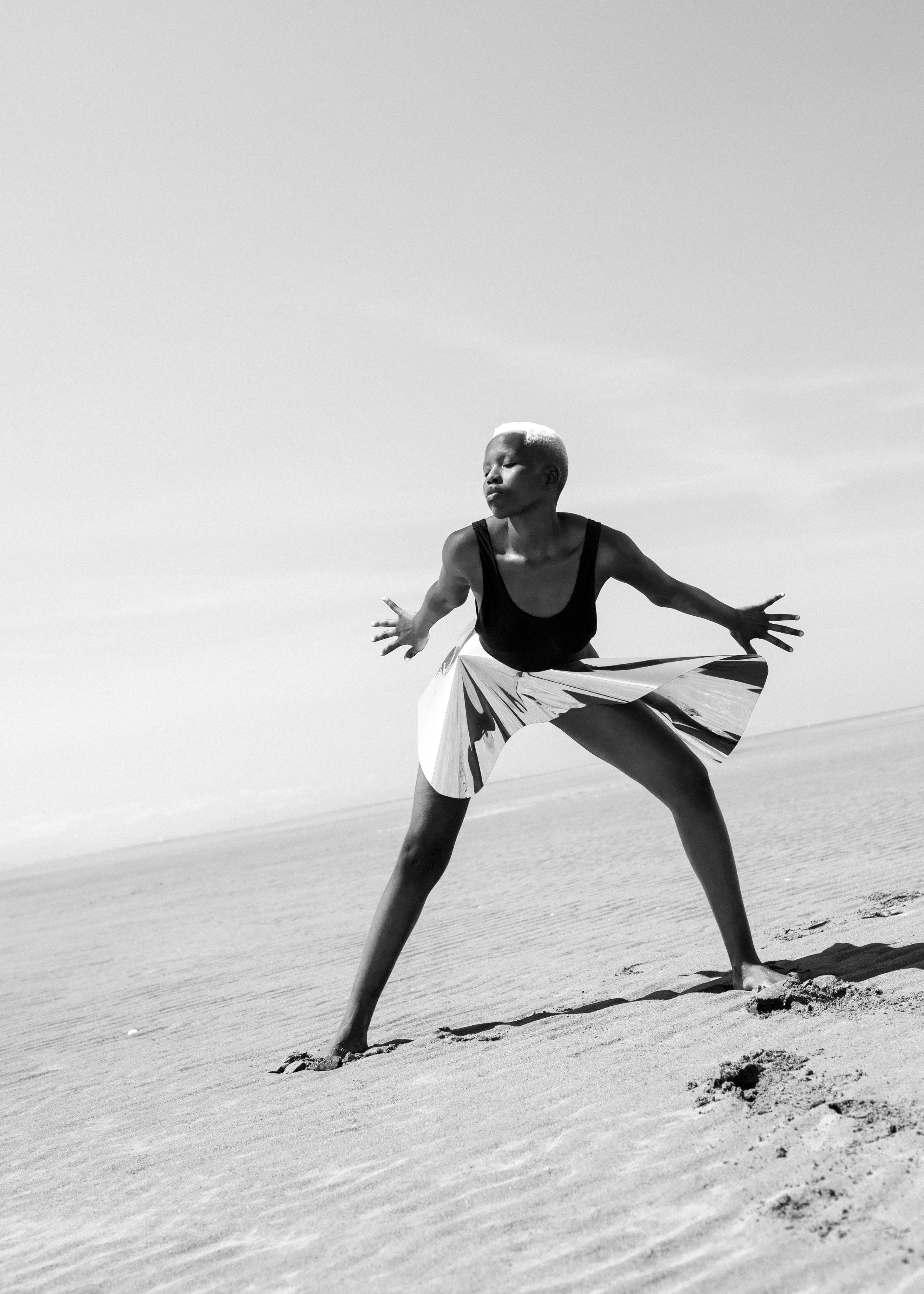 A person with a shaved head dancing or stretching on a sandy beach, wearing a black top and patterned shorts, with arms extended and eyes closed.