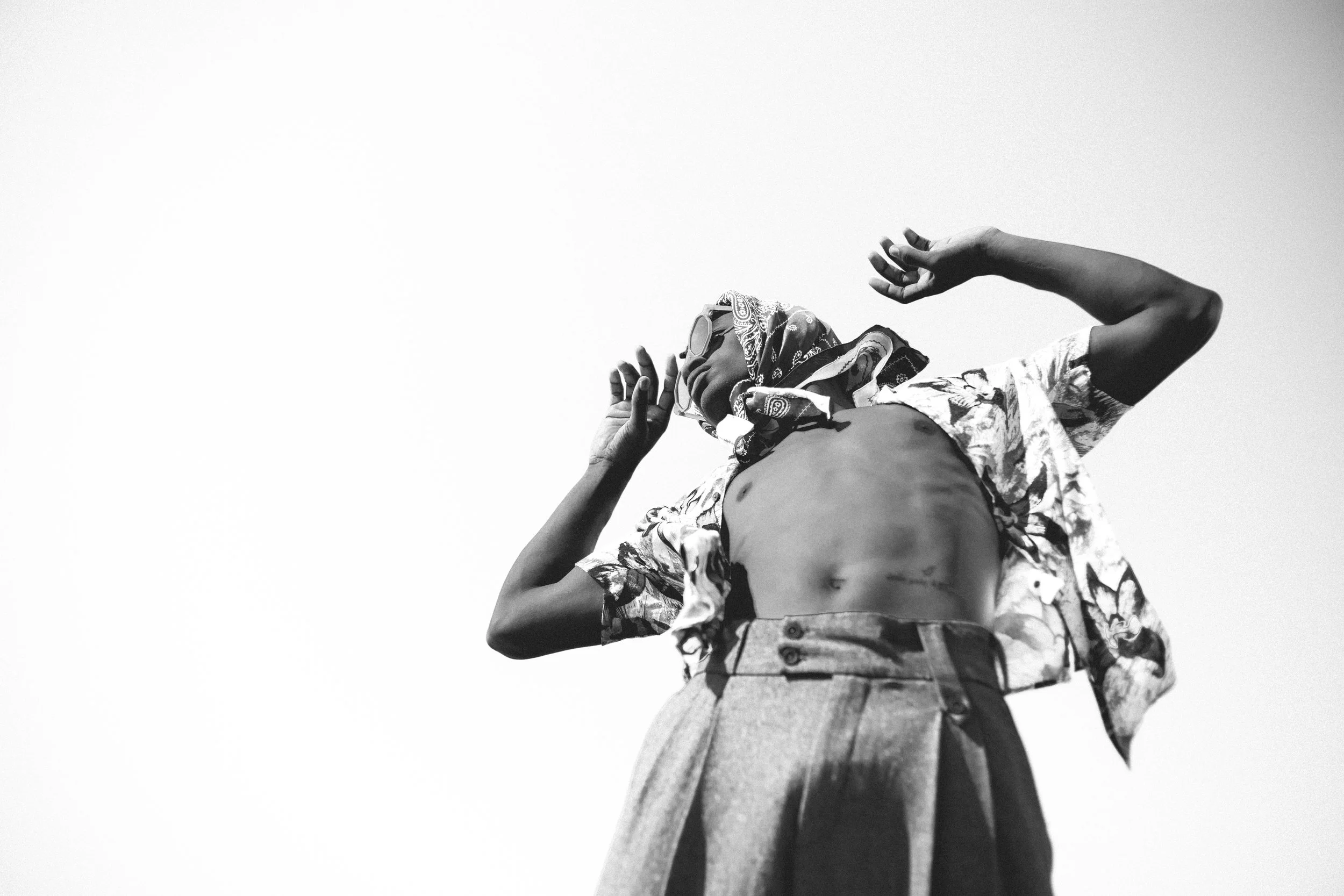 A black-and-white photo of a person outdoors, wearing sunglasses, a bandana, a floral shirt, and high-waisted pants, with arms raised and head tilted back.
