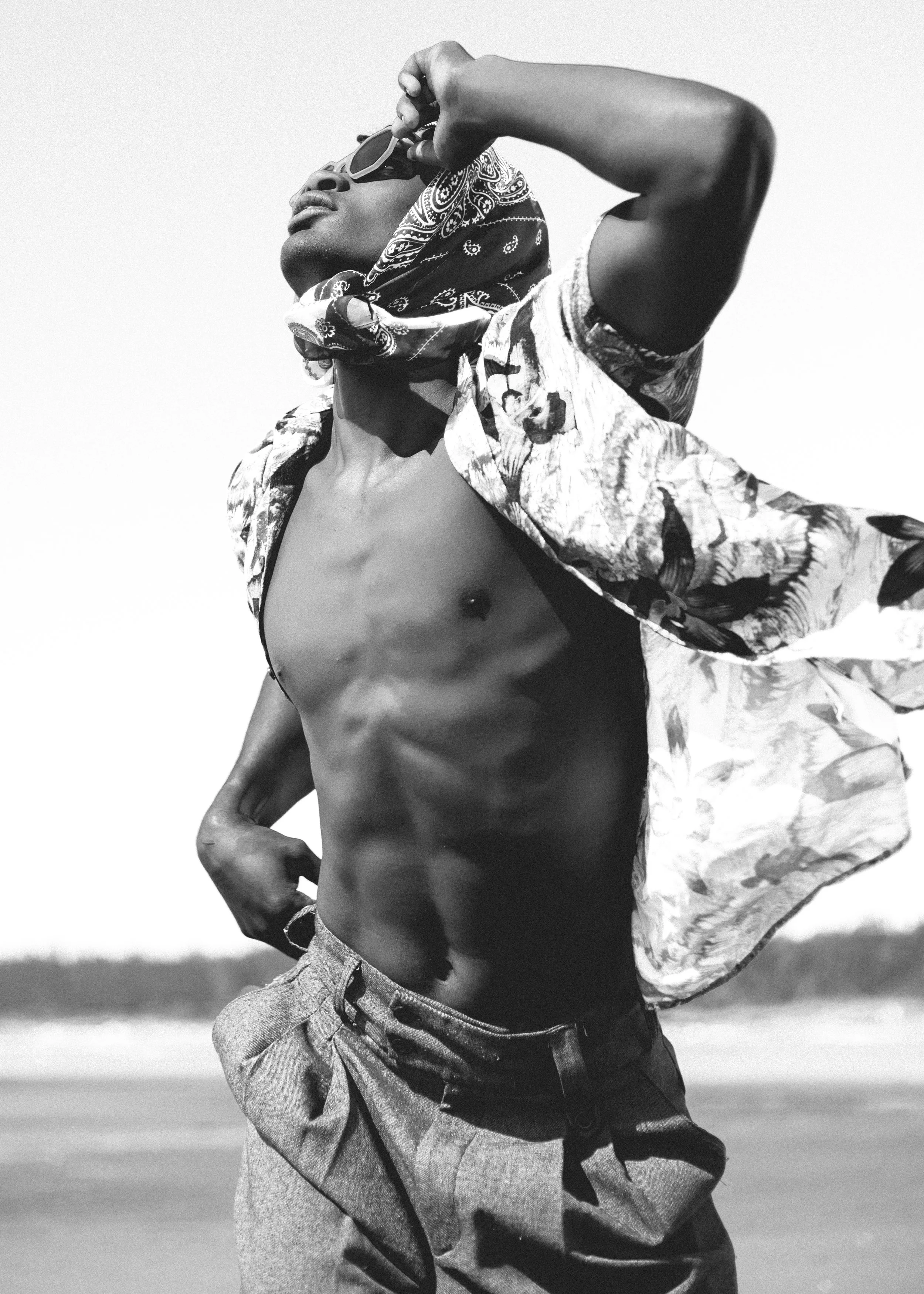 A black and white photo of a person wearing sunglasses, a bandana, a patterned shirt, and shorts, standing outdoors with their left arm raised and hand on the sunglasses.