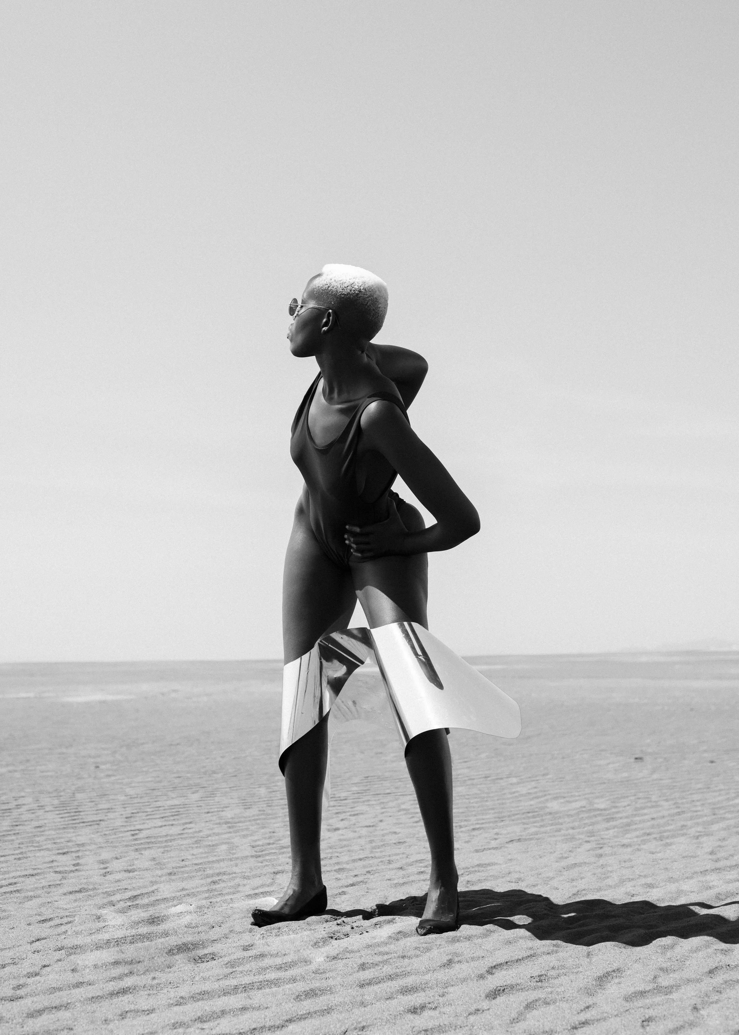 Woman with platinum blonde hair, wearing sunglasses, black top, and metallic skirt, standing barefoot on a sandy beach with a vast sky in the background.