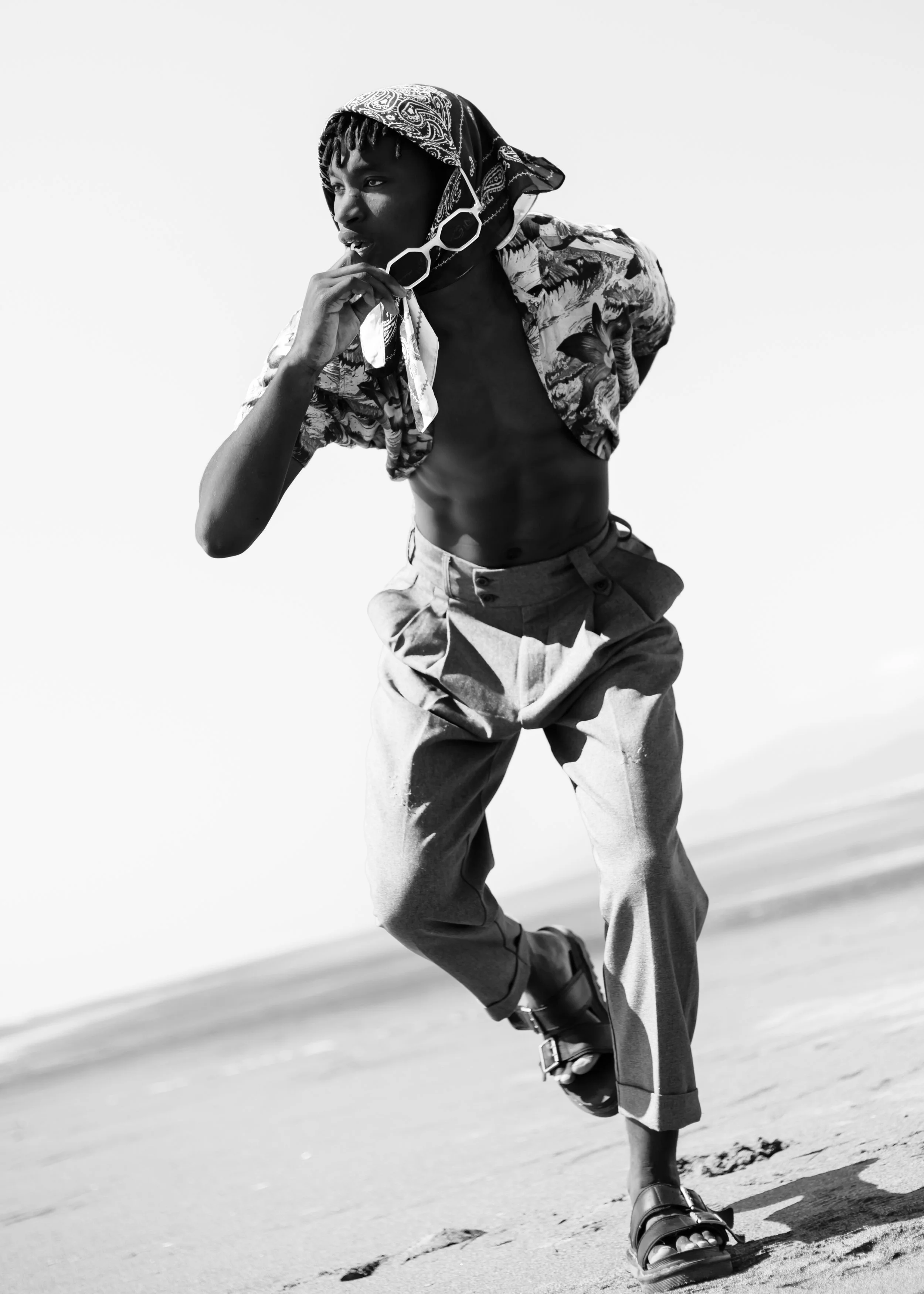 Black and white photo of a person walking barefoot on a beach, wearing loose pants, a patterned shirt, a bandana, and sunglasses, with one hand near their face.