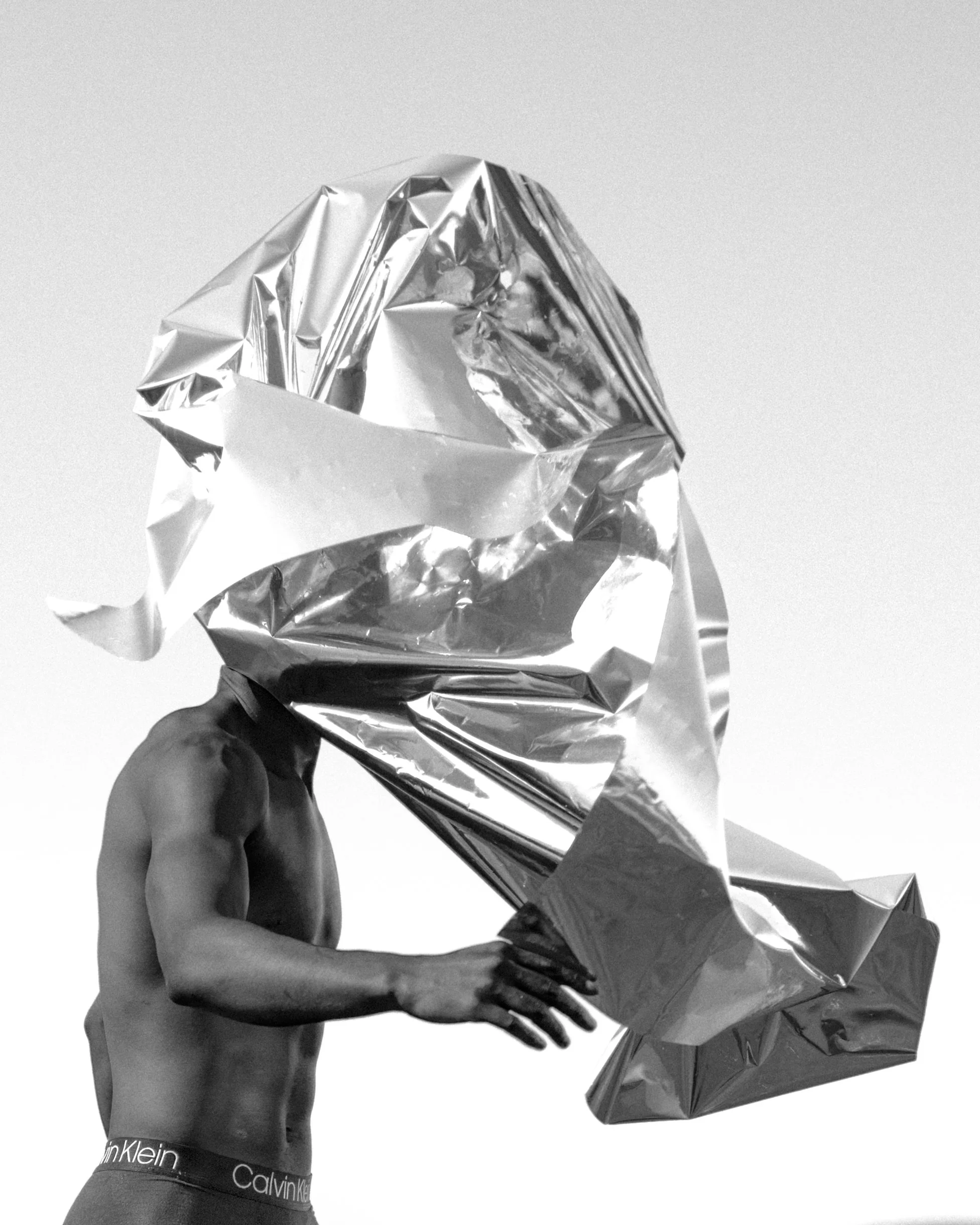 A person holding aluminum foil crumpled into a loose ball with their right hand, wearing dark Calvin Klein underwear, with no shirt, against a plain background.