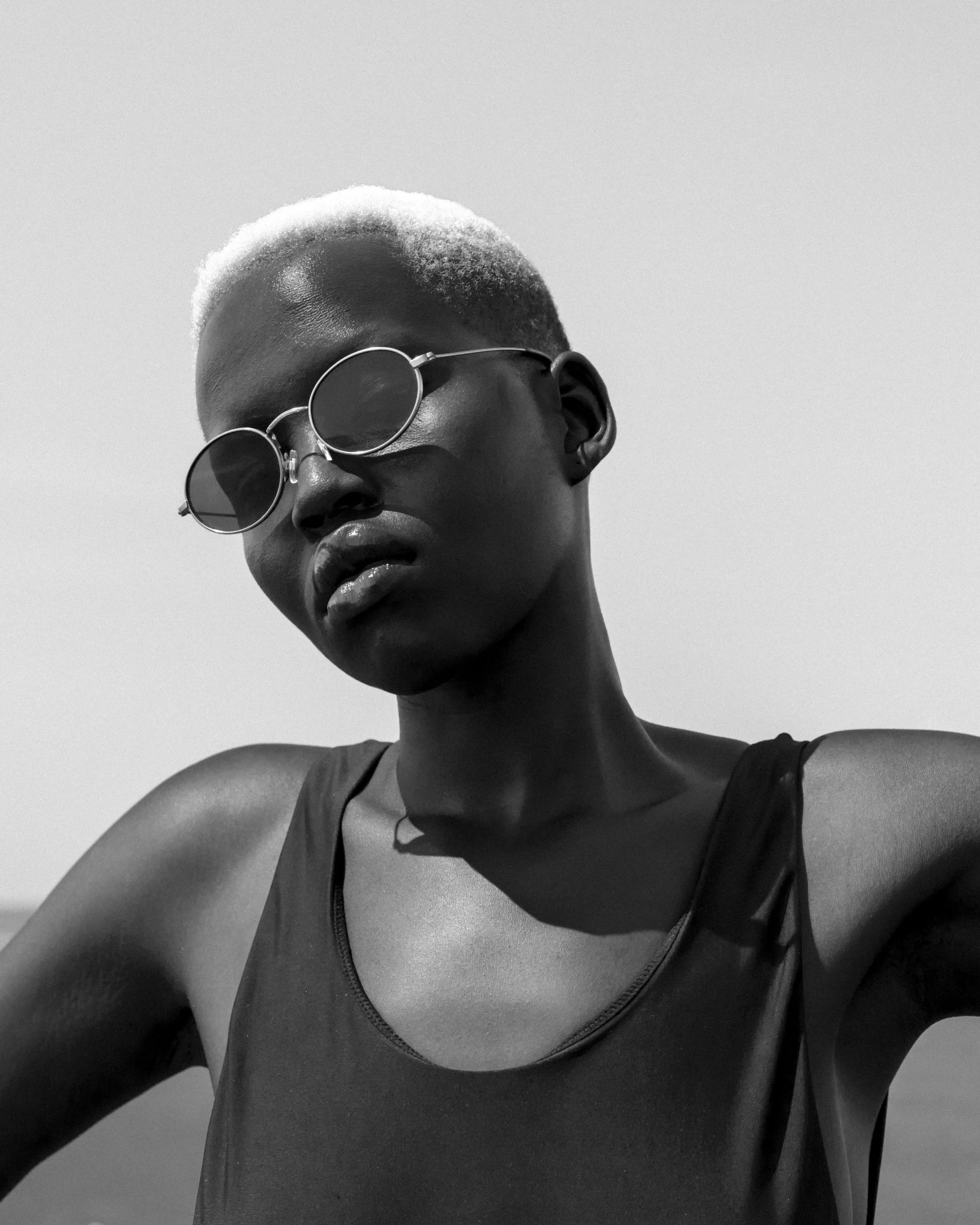 A young woman with short, platinum blonde hair wearing round sunglasses and a black tank top. She is looking slightly down, against a plain light background.