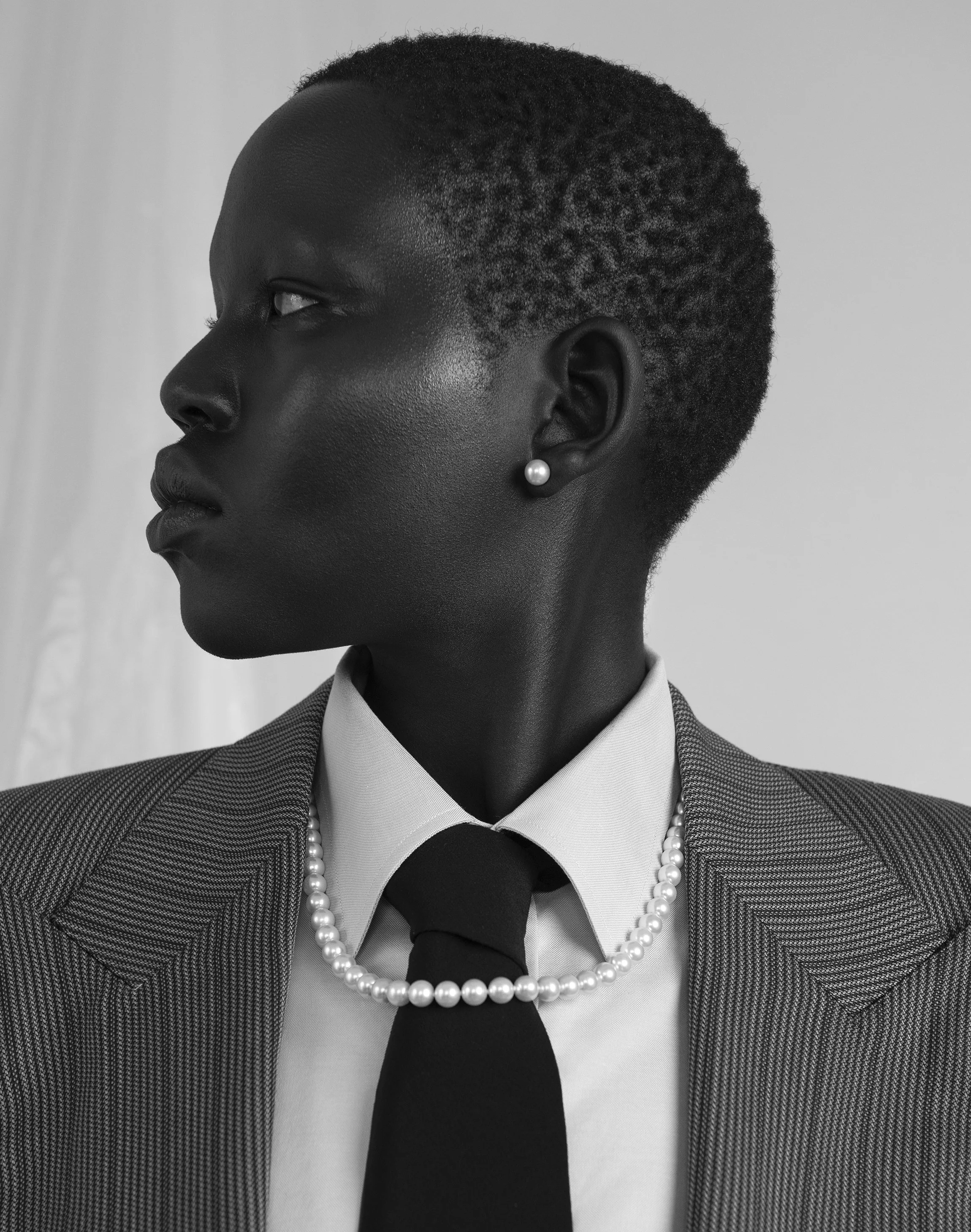 Profile portrait of a Black woman in business attire, wearing pearl jewelry, holding a microphone, in black and white.