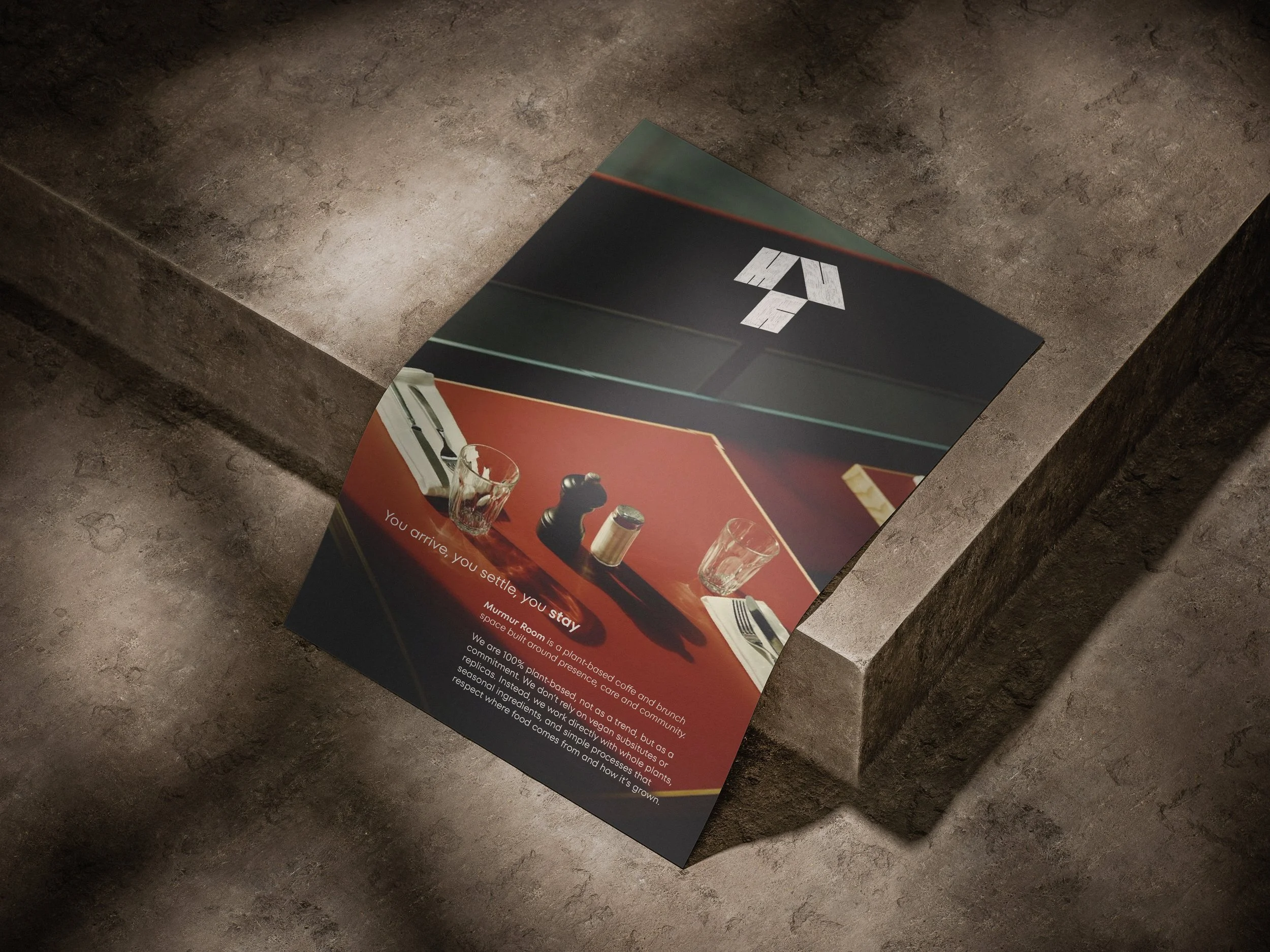 A magazine page on a dark, textured stone surface showcases a promotional ad for a plant-based coffee brand called Murmur Room. The ad features a sleek black background with white text, and an image of a glass of coffee with a spoon, a salt and pepper shaker, and a board game piece and glasses on a red surface. The headline reads 'You arrive, you settle, you stay,' emphasizing the brand's focus on natural ingredients and environmental sustainability.