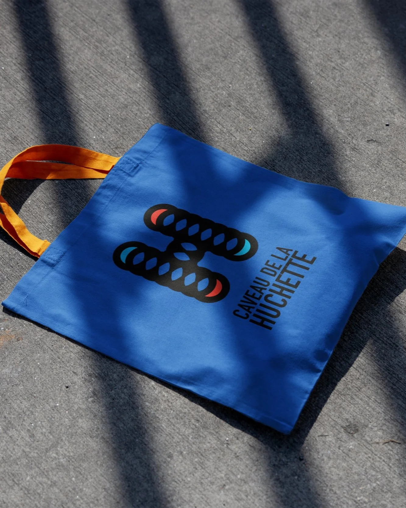 A blue reusable shopping bag with orange handles, displaying a black and red hatching tape symbol, and the text 'CAVEAU DE LA HUILE' printed below it, on a concrete surface with shadows.