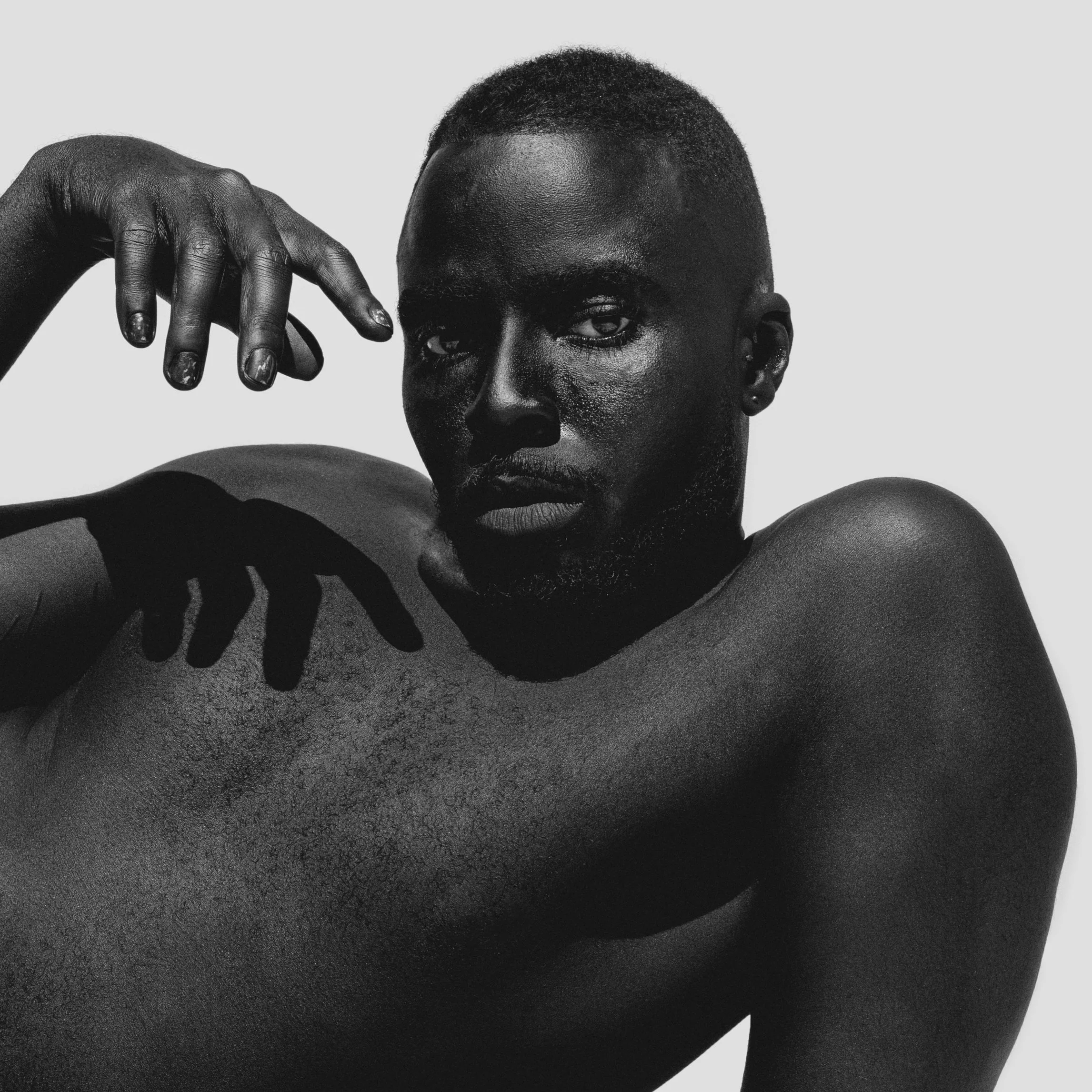 Black and white close-up of a person with short hair and dark skin, resting their hand on the side of their face and arm, against a plain light gray background.