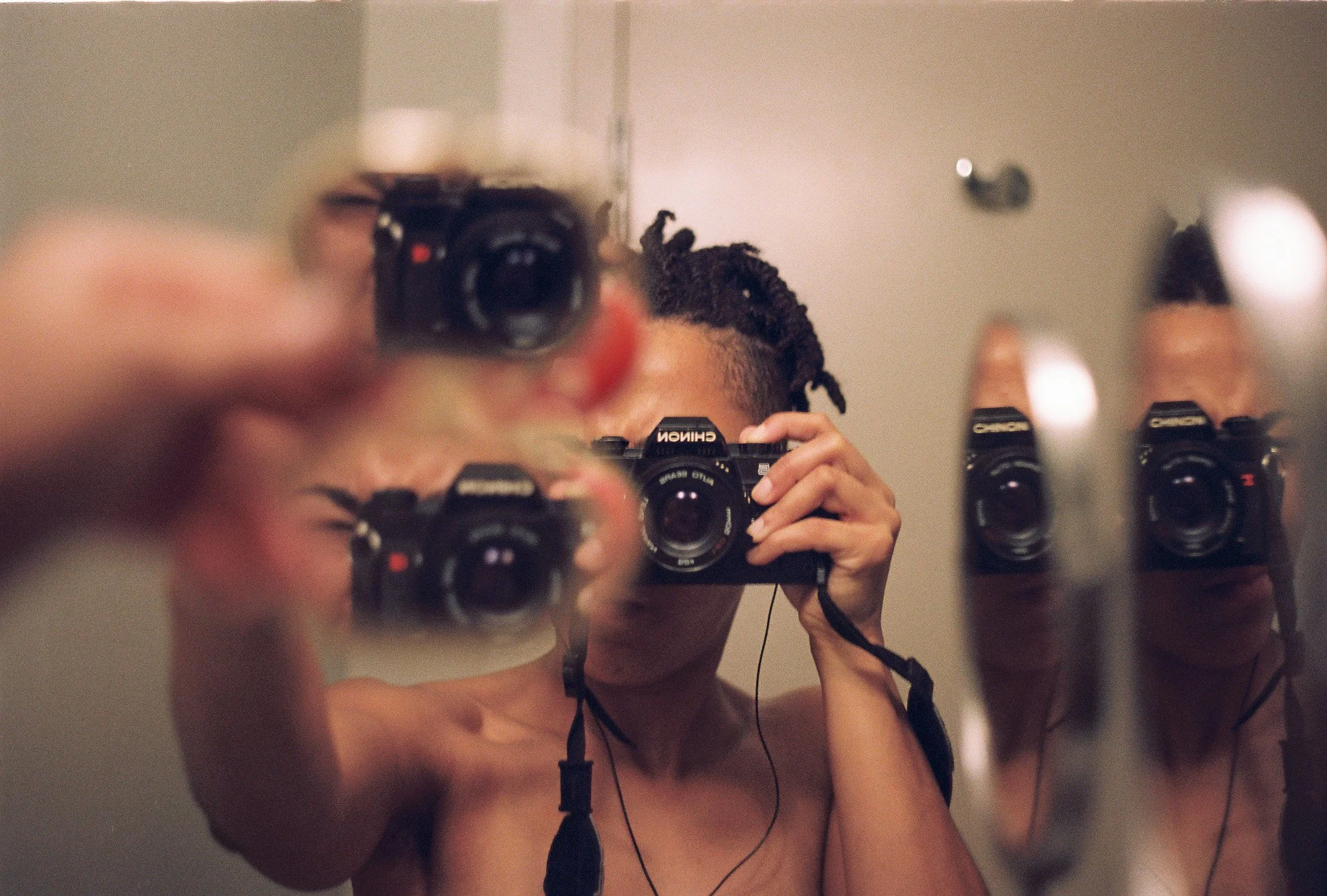 a photographer taking a photograph of themself using a handheld mirror and large mirror creating a multiplication of themself in the mirrors