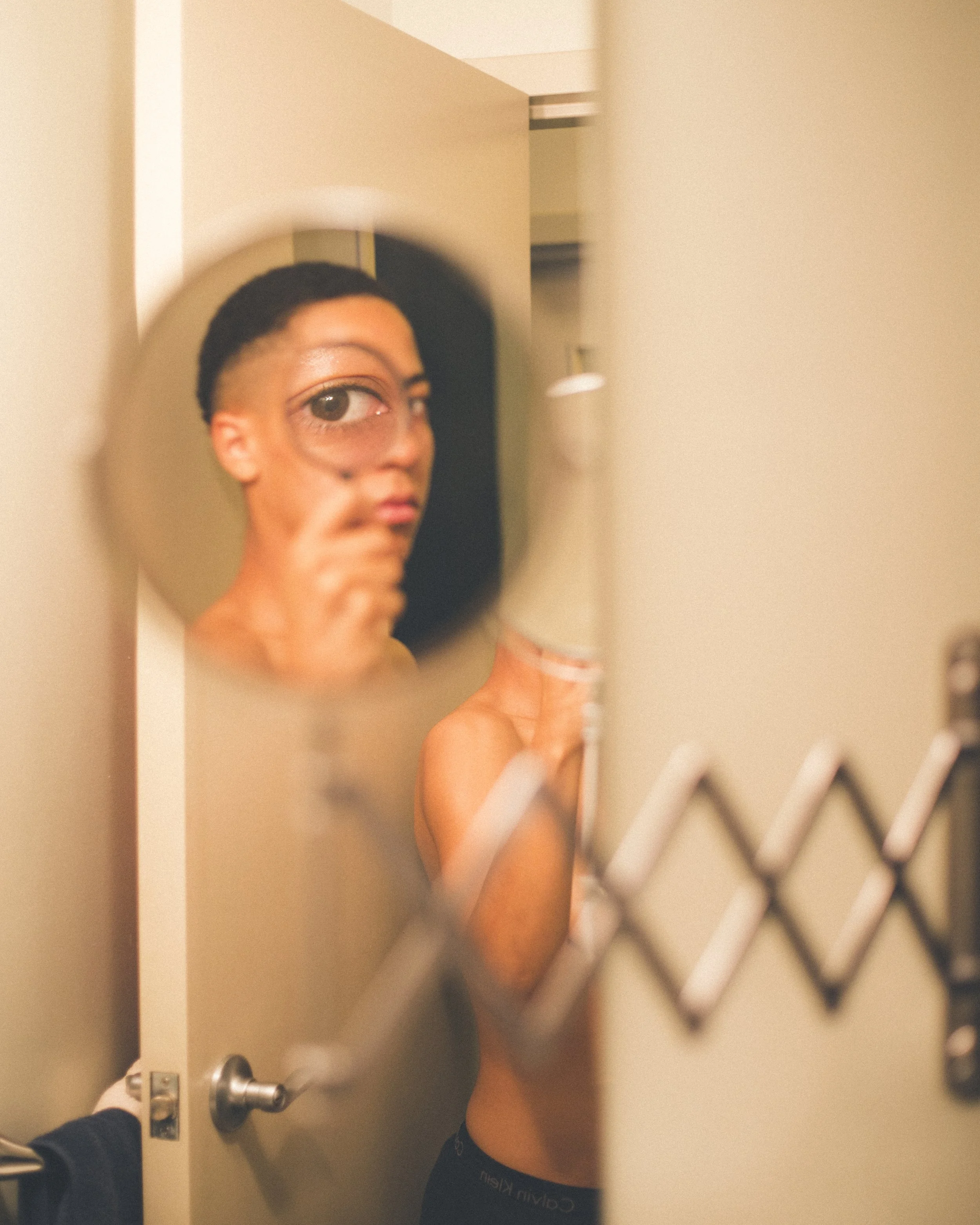 person taking a photo of themselves using a mirror and magnifier with their body half naked and halfway through a door