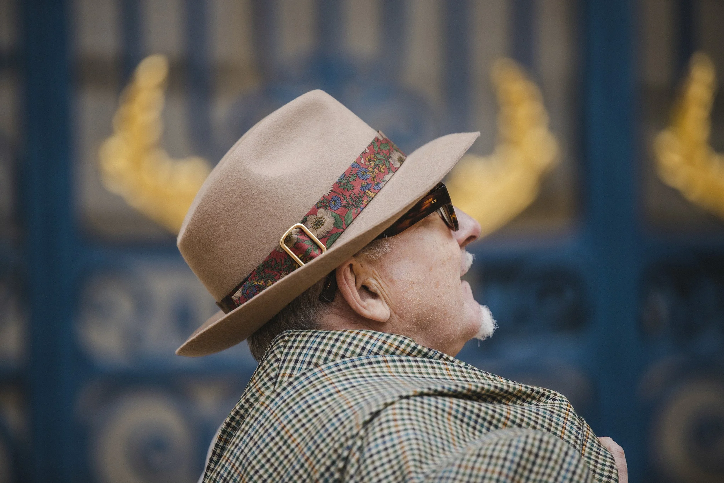 A close-up profile of an elderly man wearing a beige felt fedora hat with a colorful floral ribbon, sunglasses, and a plaid jacket, standing outdoors with a blurred background of blue and yellow elements.