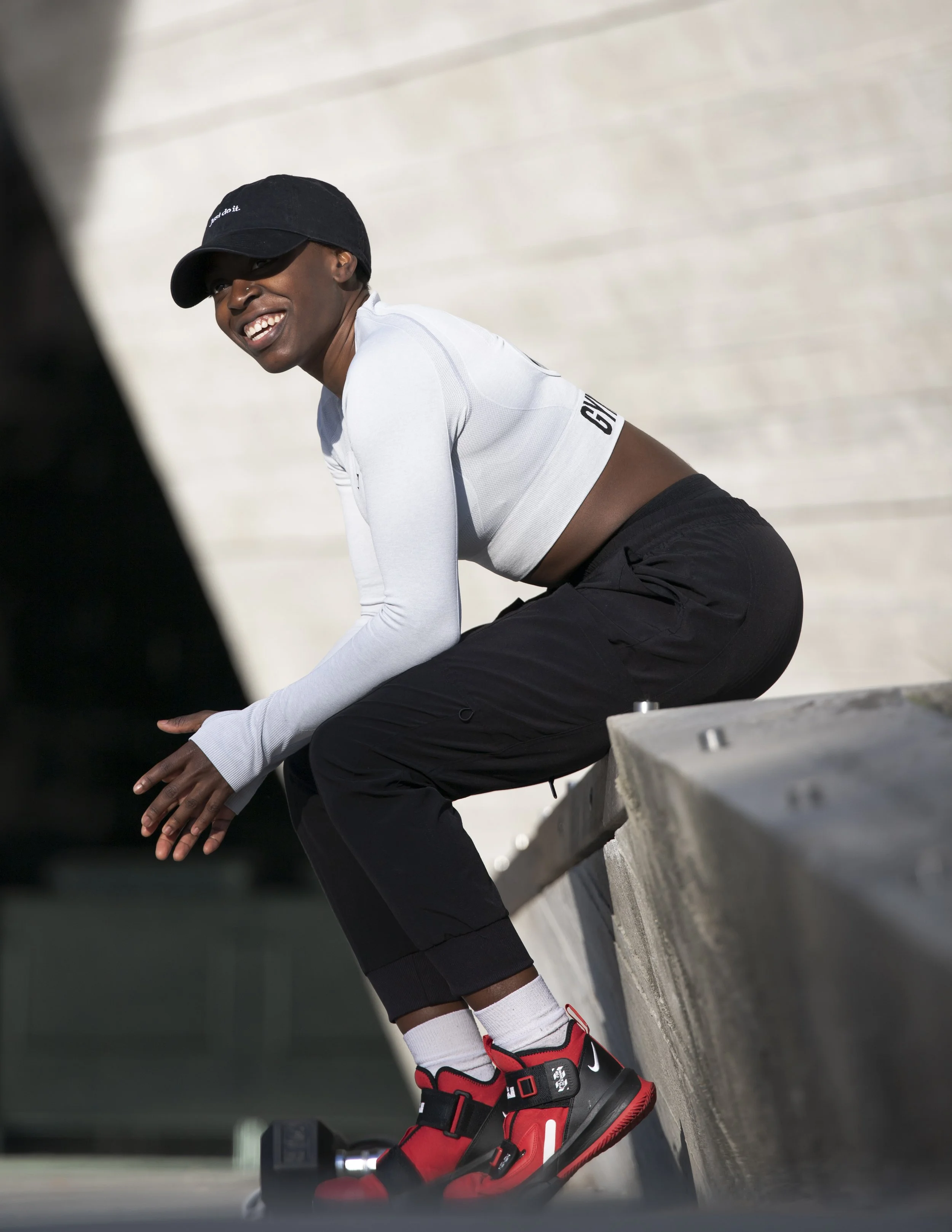 A person wearing a black cap, white long-sleeve shirt, and black shorts is kneeling on a concrete surface. They are smiling and looking to the side. The person is wearing red and black high-top sneakers and white socks with a logo. The background is 