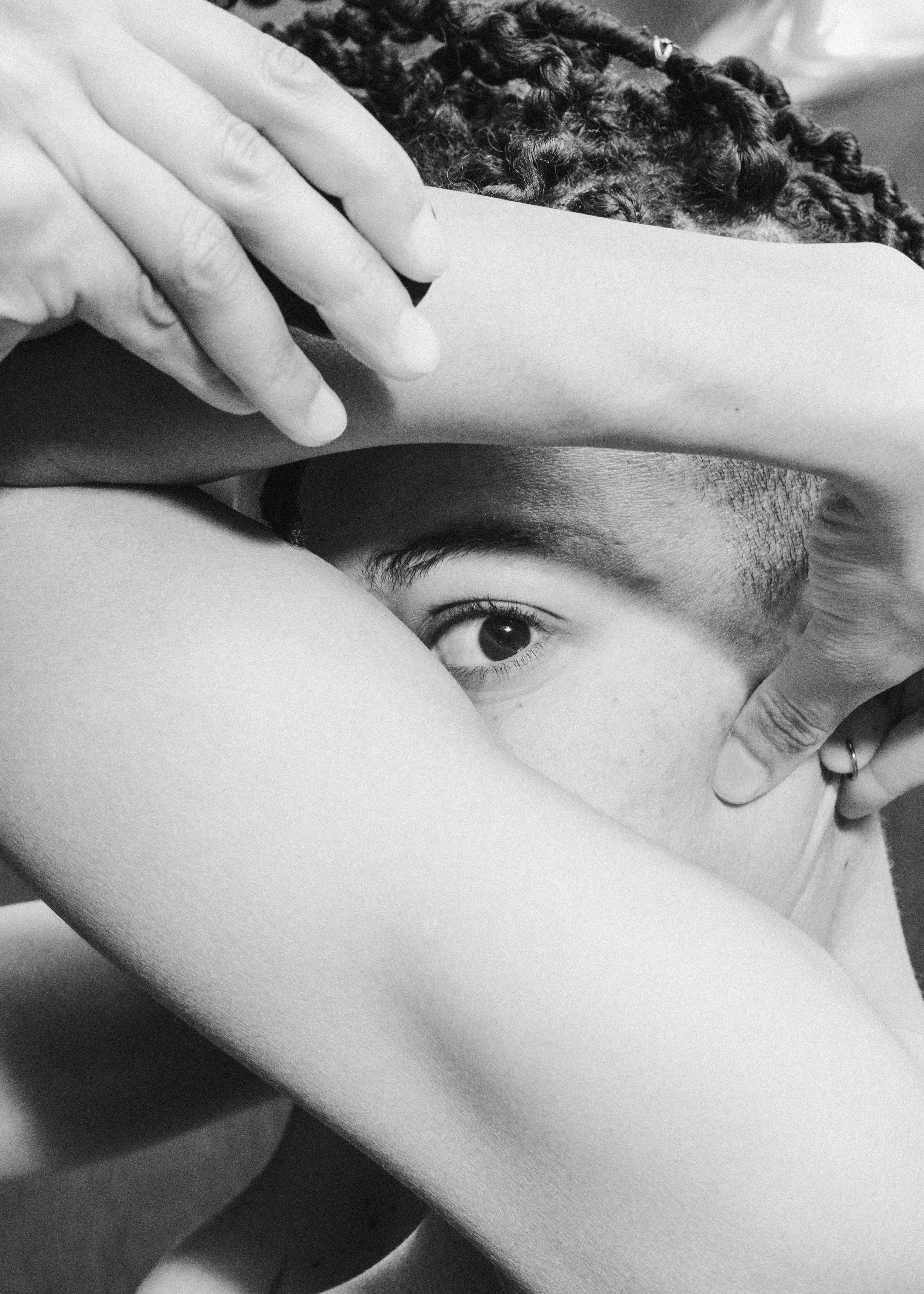 A person's eye peeking out from beneath their arm, with their hand resting on their face, and another person's hand gently touching their arm.